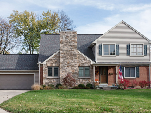 Two-story house with stone chimney, brick and beige siding, green lawn, garage, and blue sky.