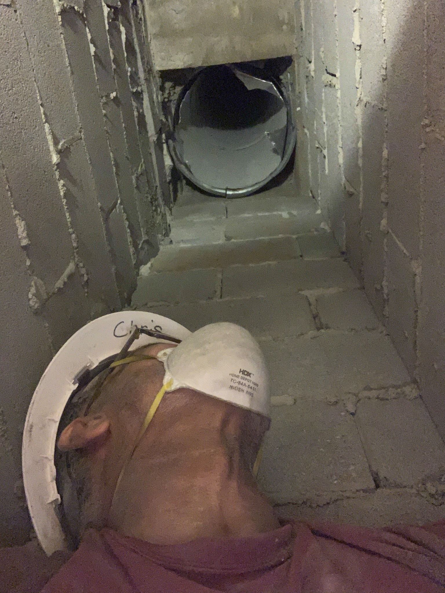 Person in hard hat and mask looking into a dark duct within a concrete block structure.