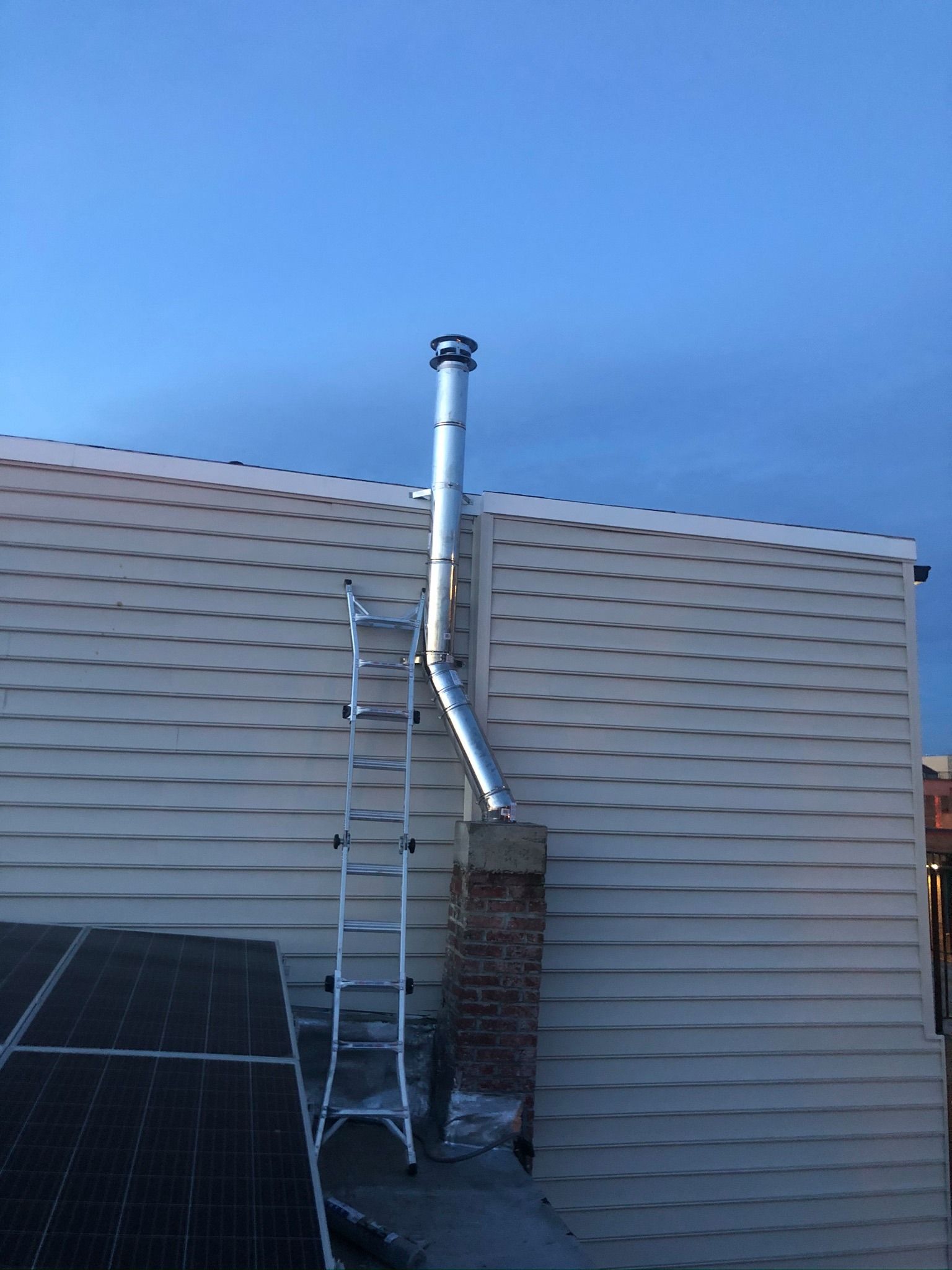 A metal chimney with a ladder on a beige siding wall next to solar panels and blue sky.