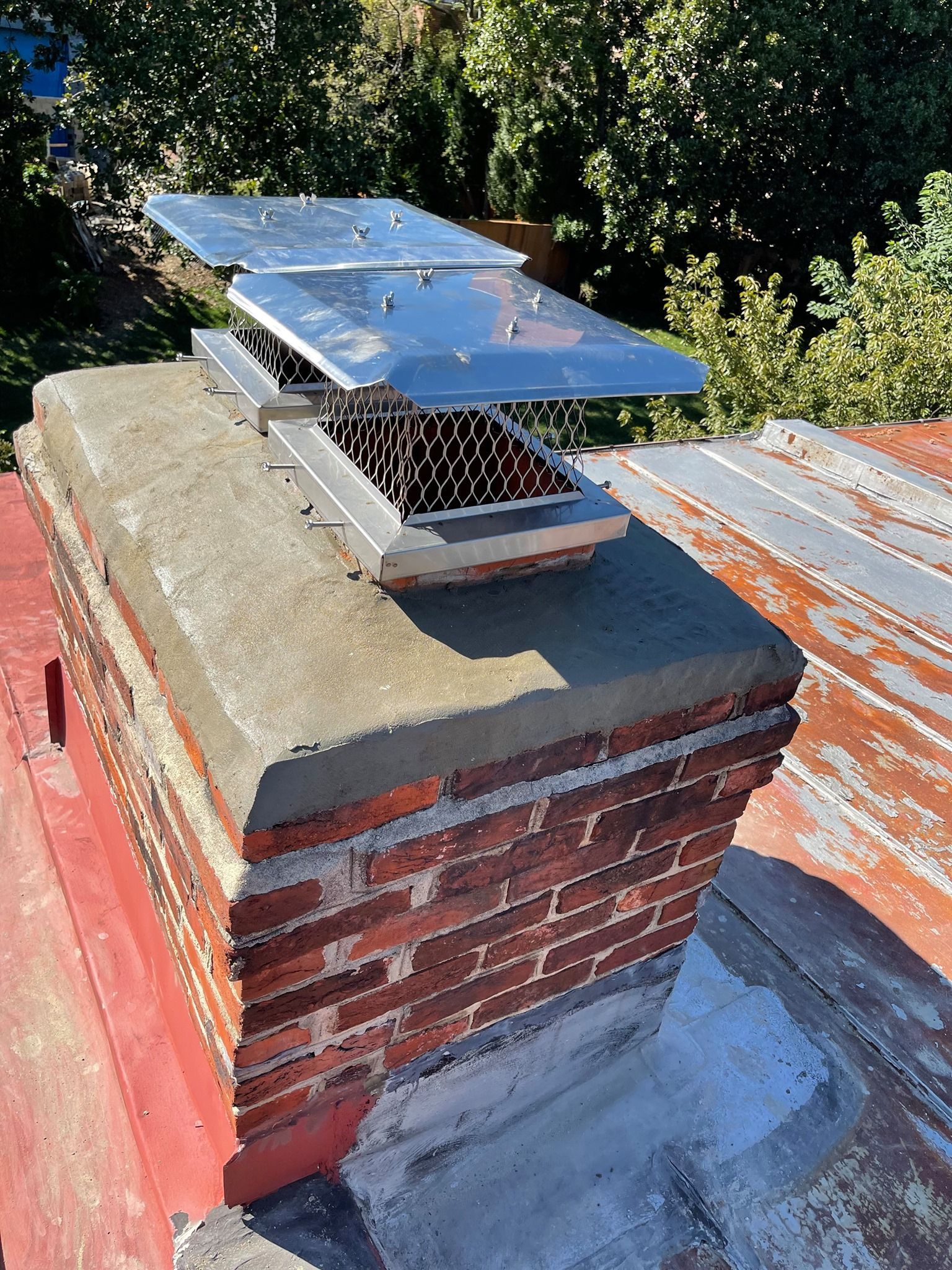 Brick chimney with metal cap and top, on a red rooftop, surrounded by greenery.