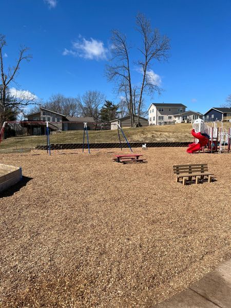 Playground with swings, slides, picnic table, bench, and wood chip ground. Houses in background.