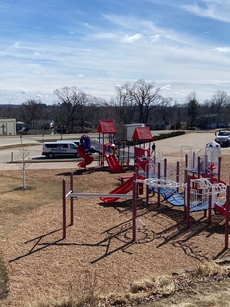 Playground with red, white, and blue structures on wood chip ground. Trees, van, and blue sky in background.