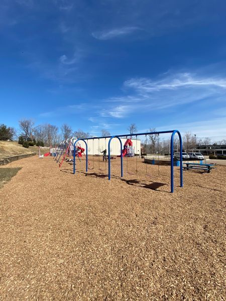 Playground with blue swing sets and red swings, on a wood chip surface, under a blue sky.