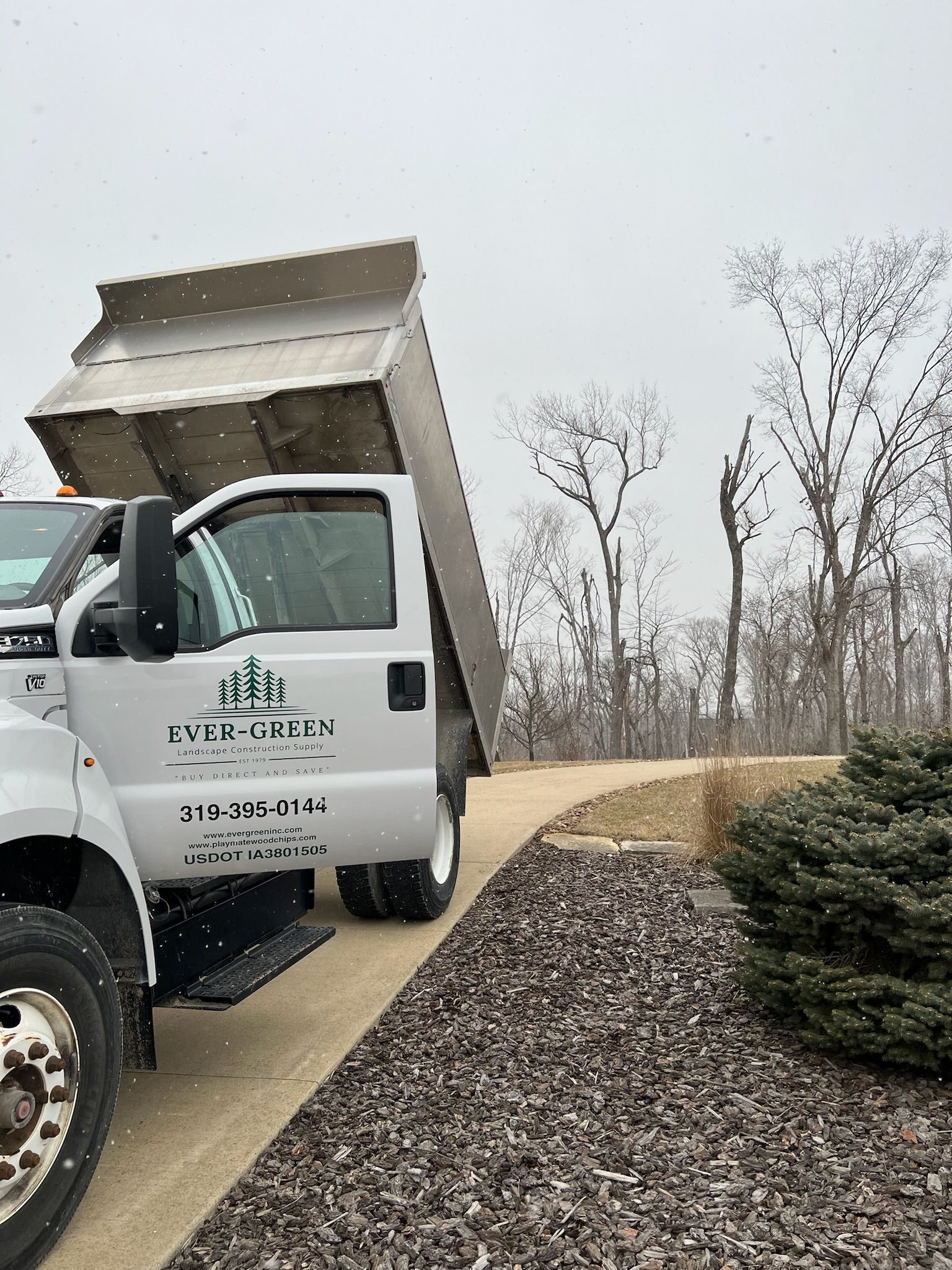 White dump truck unloading gravel on a snowy day; landscaping business logo on the door.