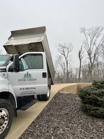 White dump truck unloading gravel on a snowy day; landscaping business logo on the door.