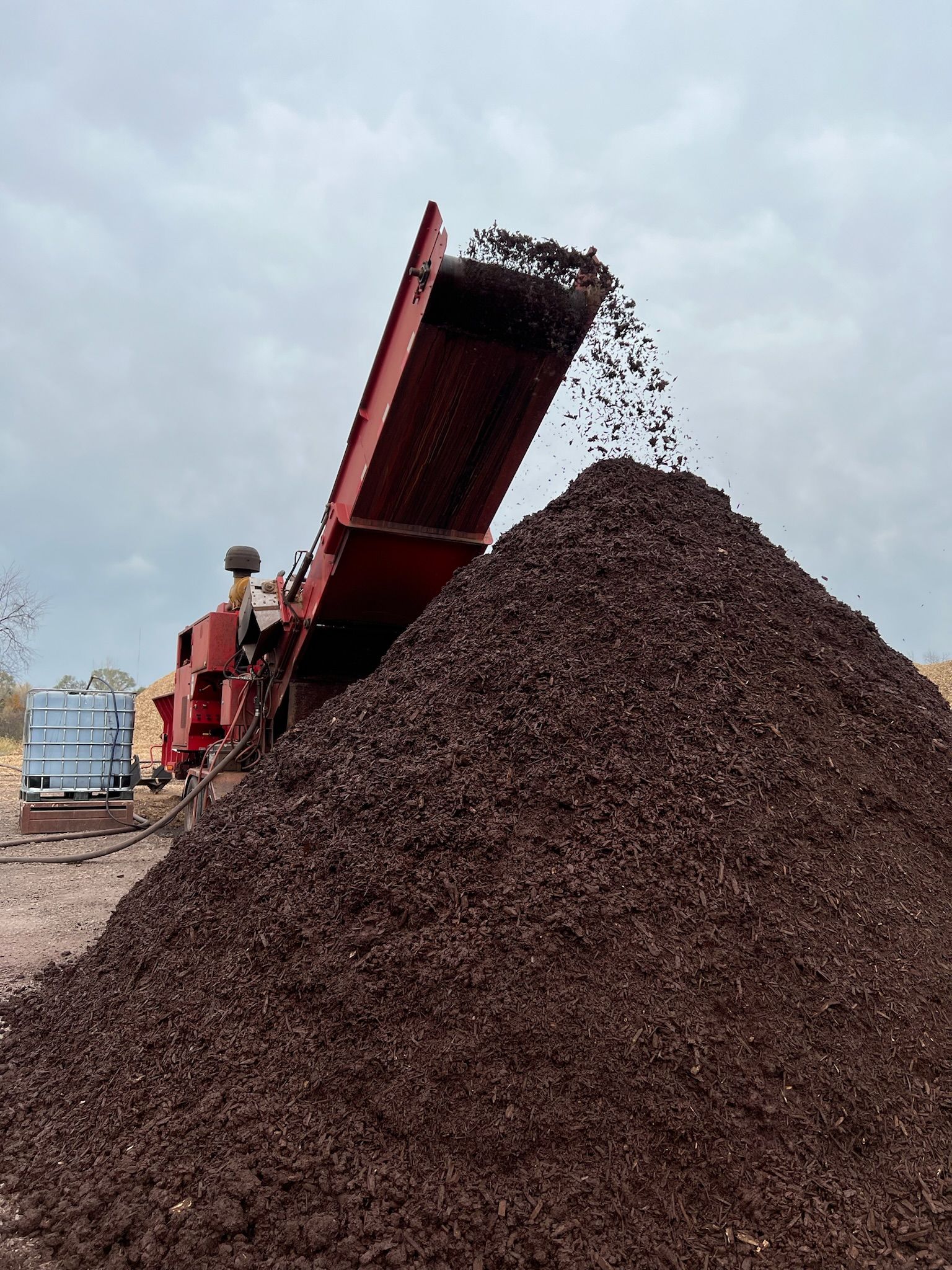 Dark mulch being discharged from a red conveyor belt into a large pile. Cloudy sky.