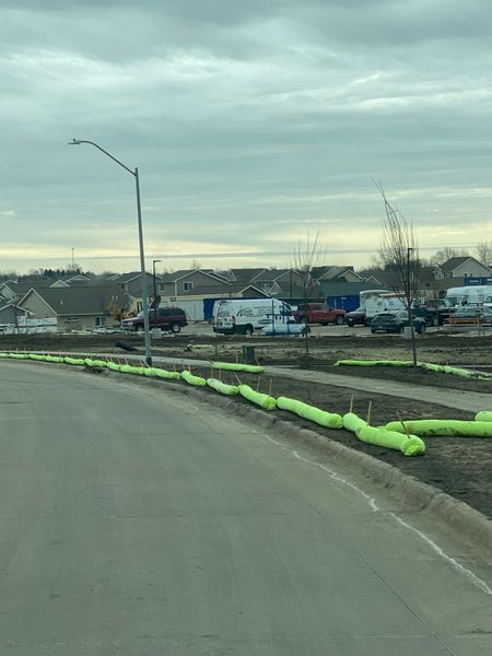 Road with green sediment control tubes, construction site in background with vehicles and houses. Overcast sky.