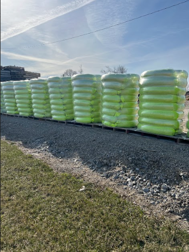 Green-wrapped bales of material stacked in a row on a gravel surface outdoors under a blue sky.