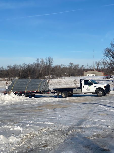 White truck towing a flatbed trailer loaded with metal pipes in a snow-covered parking lot on a sunny day.