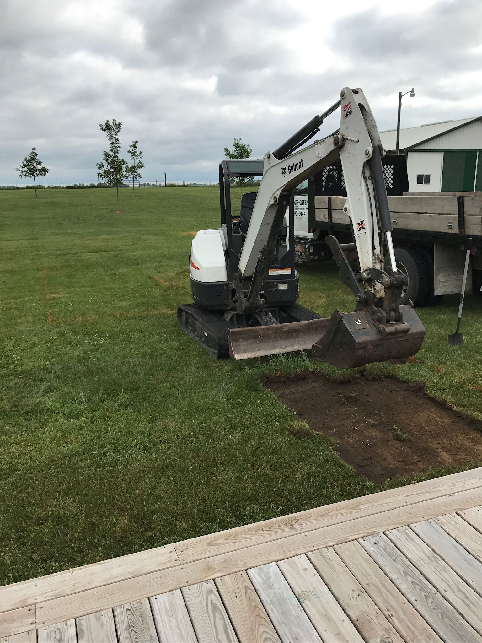 Mini excavator digging a trench in a grassy area next to a wooden deck. Cloudy sky.