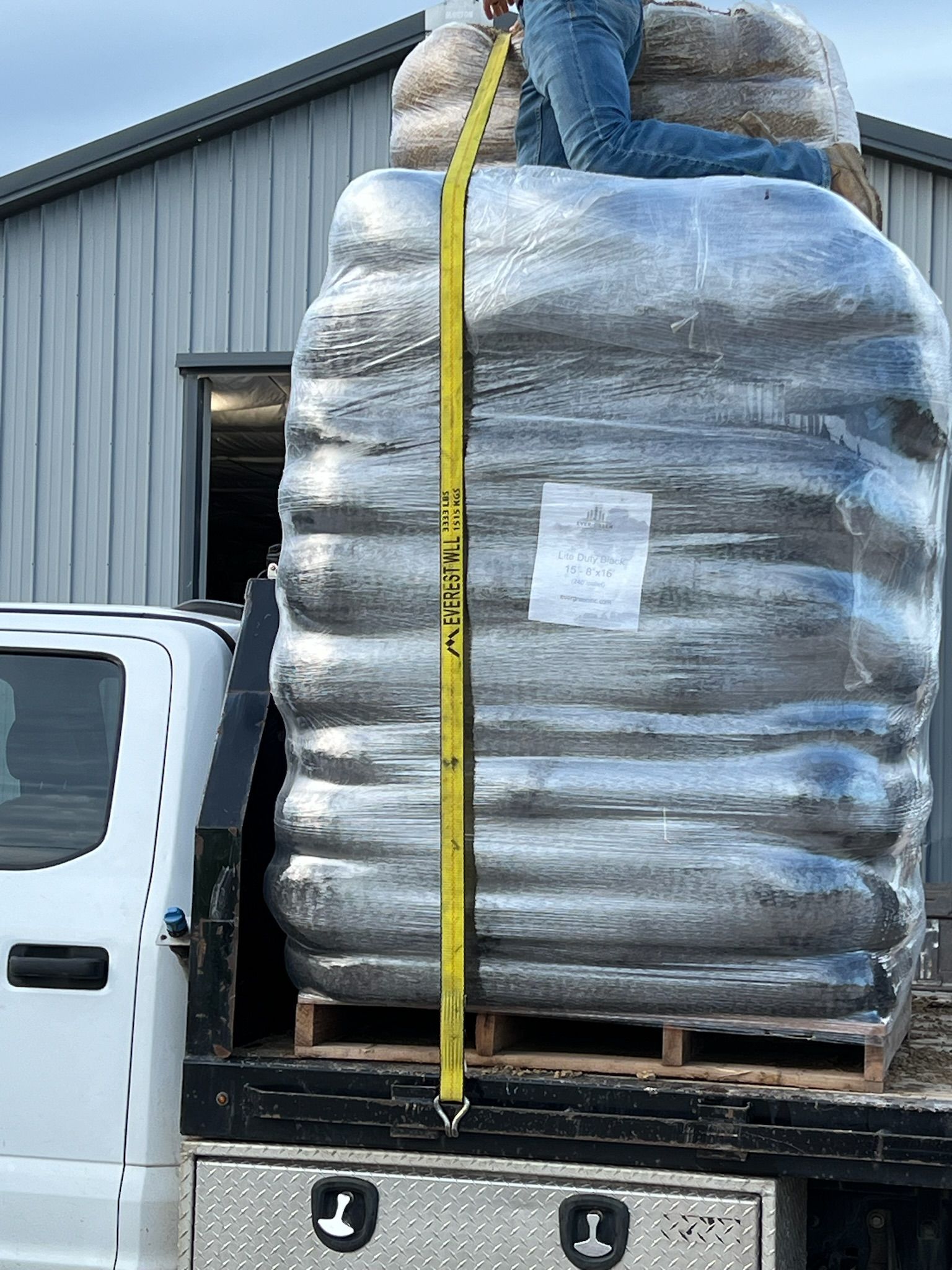 Person securing a large, wrapped pallet of materials on a truck bed with a yellow strap.