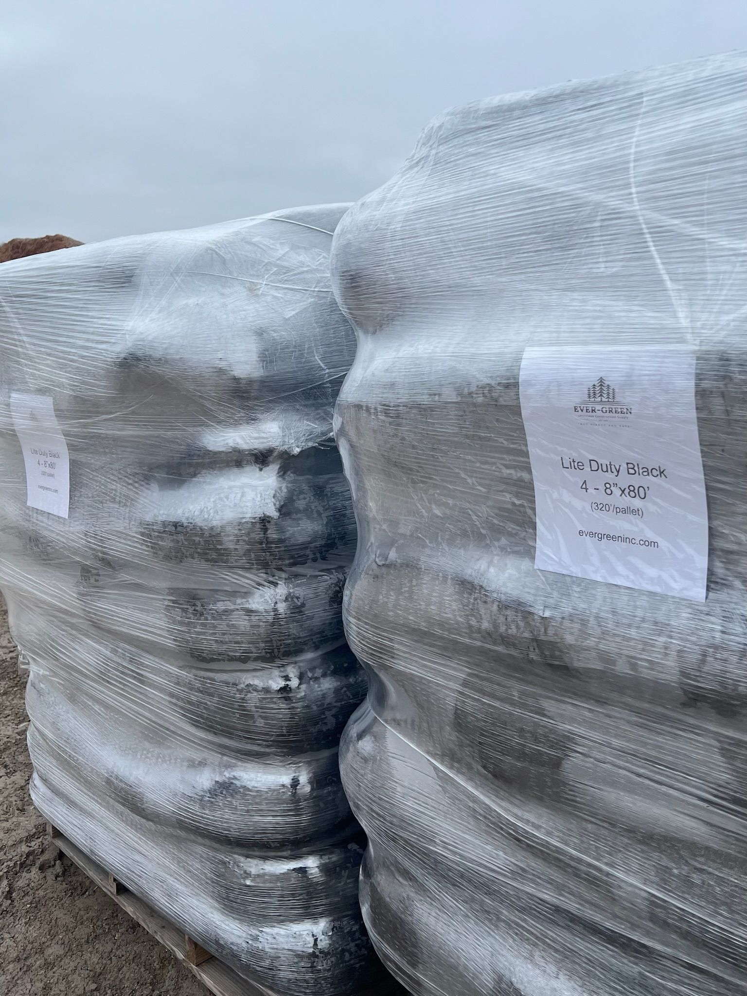 Two wrapped pallets of dark-colored material outdoors, likely for construction. White labels are attached.