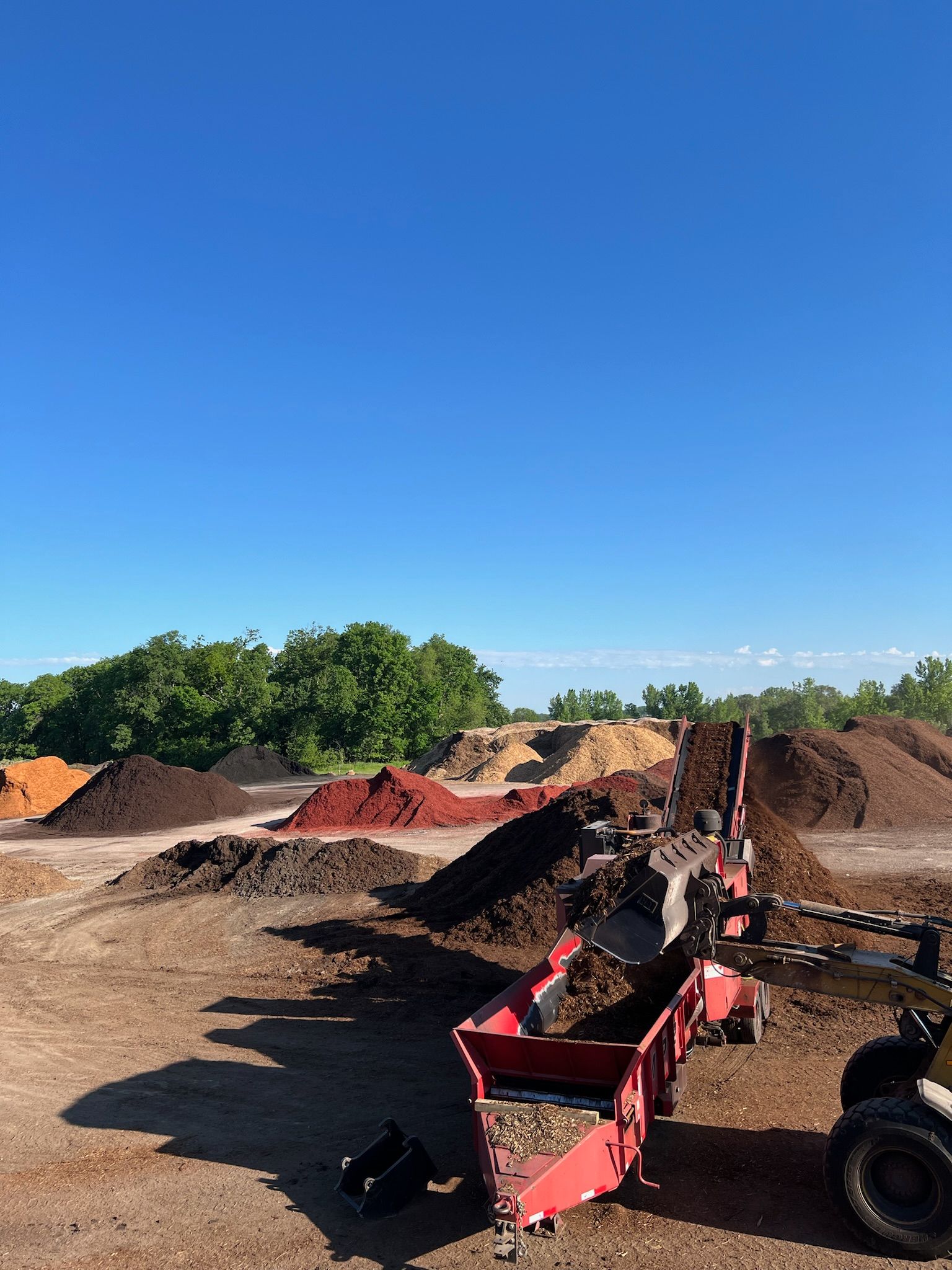 Mulch screening machine in a yard with piles of various colored mulch under a clear blue sky.