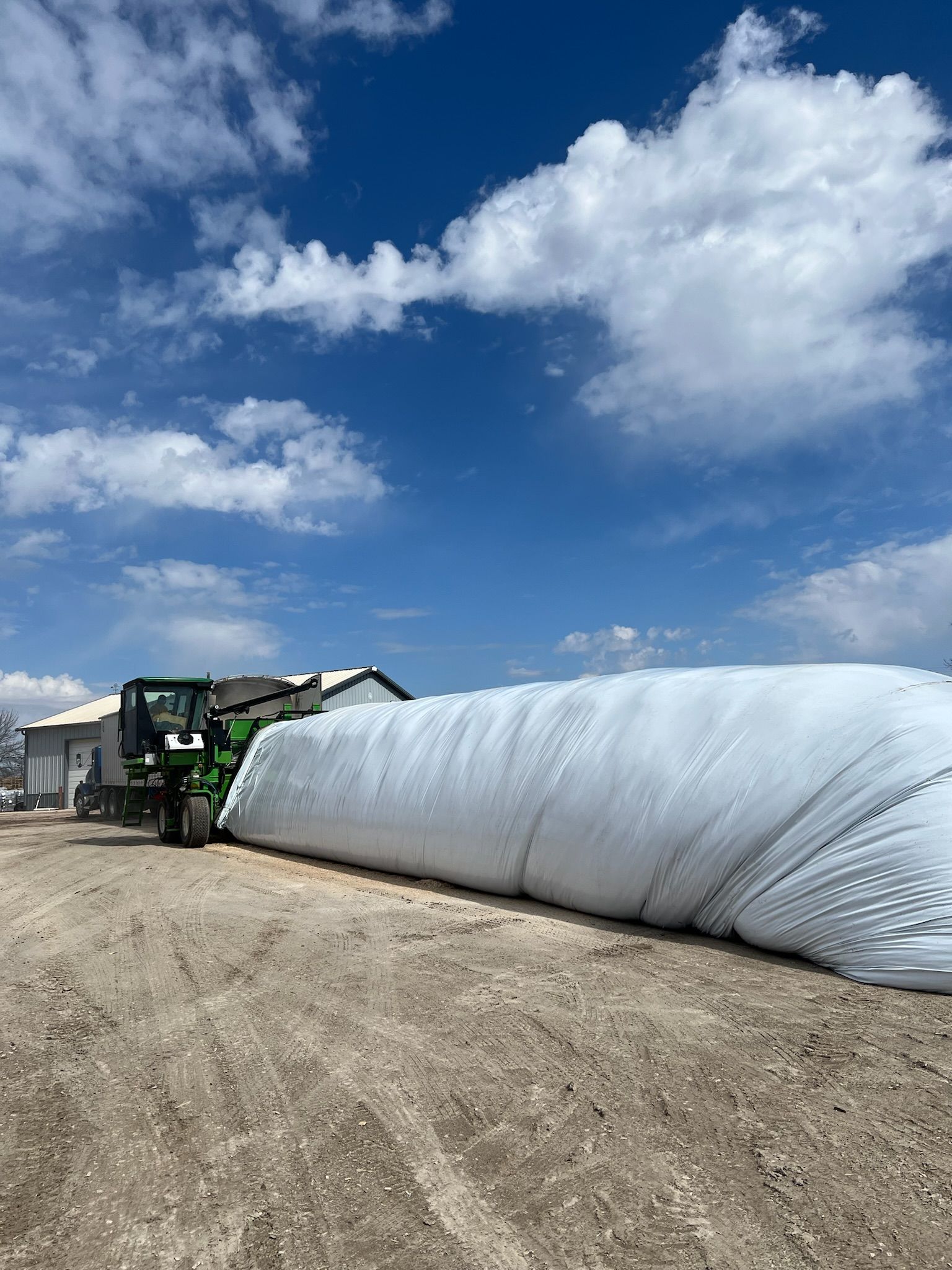A tractor next to a white silage storage bag on a dusty farm under a blue sky with clouds.