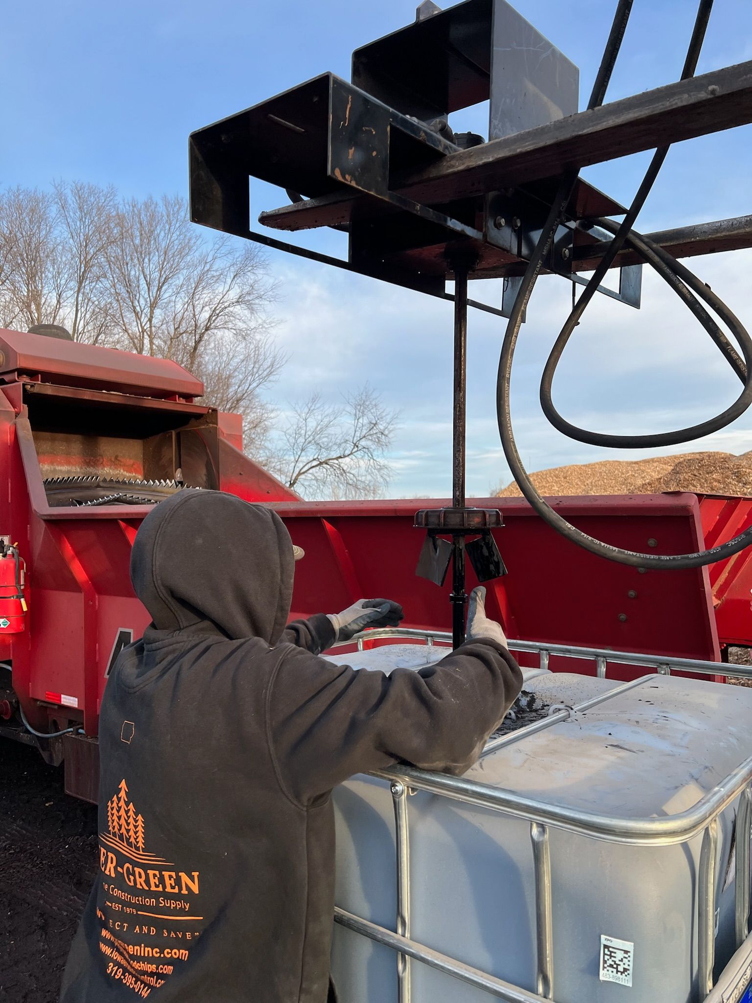Person in a hoodie operating machinery to fill a large white container, red machine in the background.