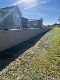 Long concrete wall alongside a grassy area, next to a house and shed on a sunny day.