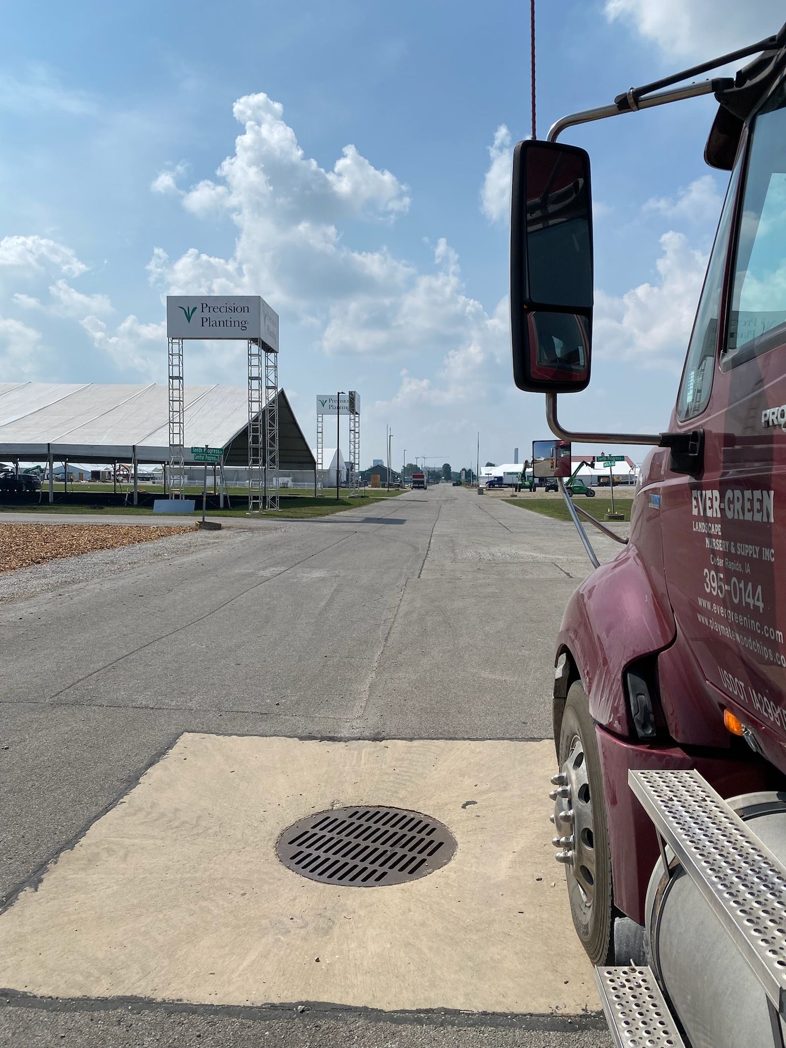 A semi-truck parked on a gray road, with a manhole cover in front. Buildings and blue sky in the background.