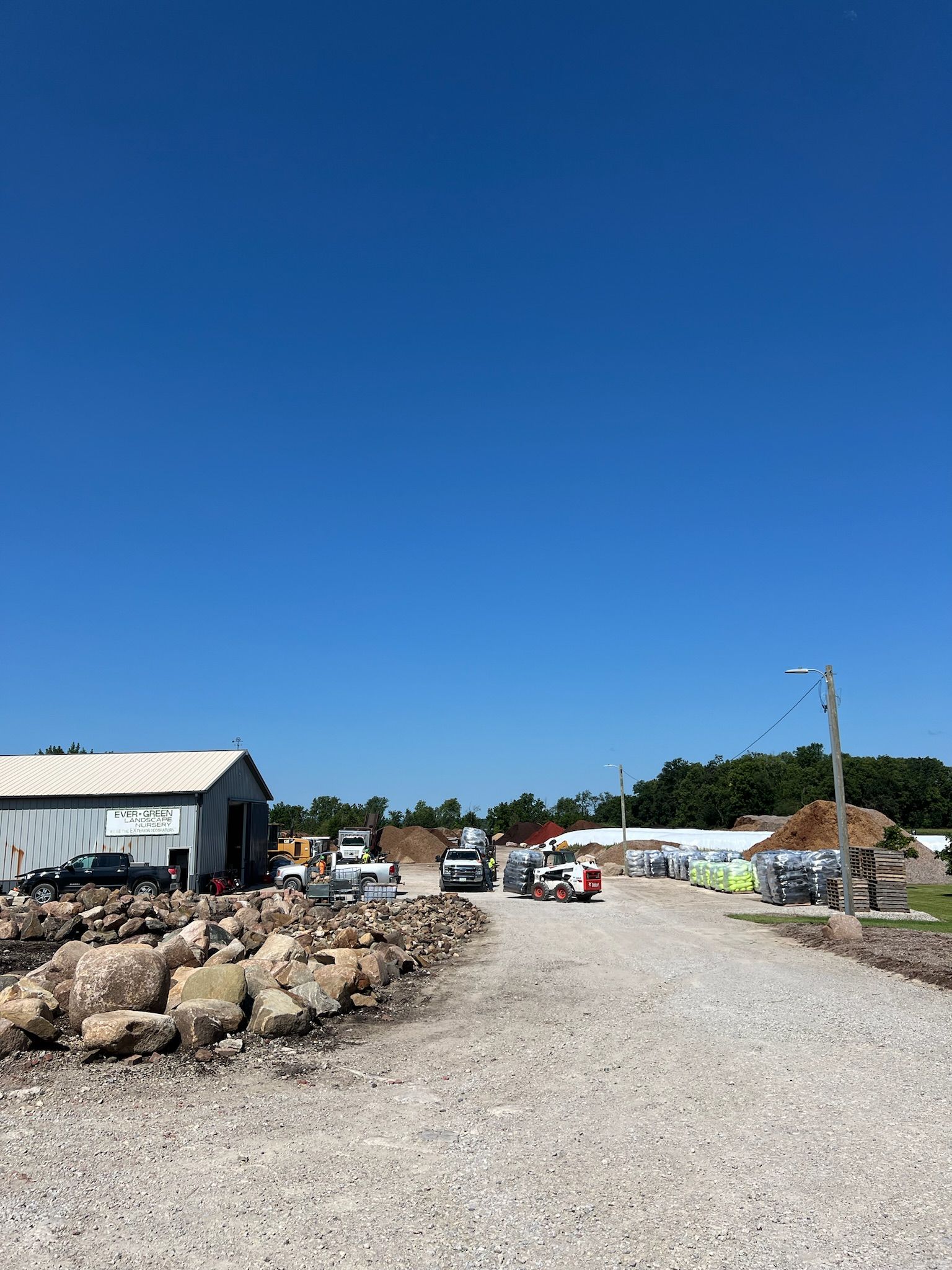 A gravel road leads past buildings, vehicles, and construction materials under a blue sky.