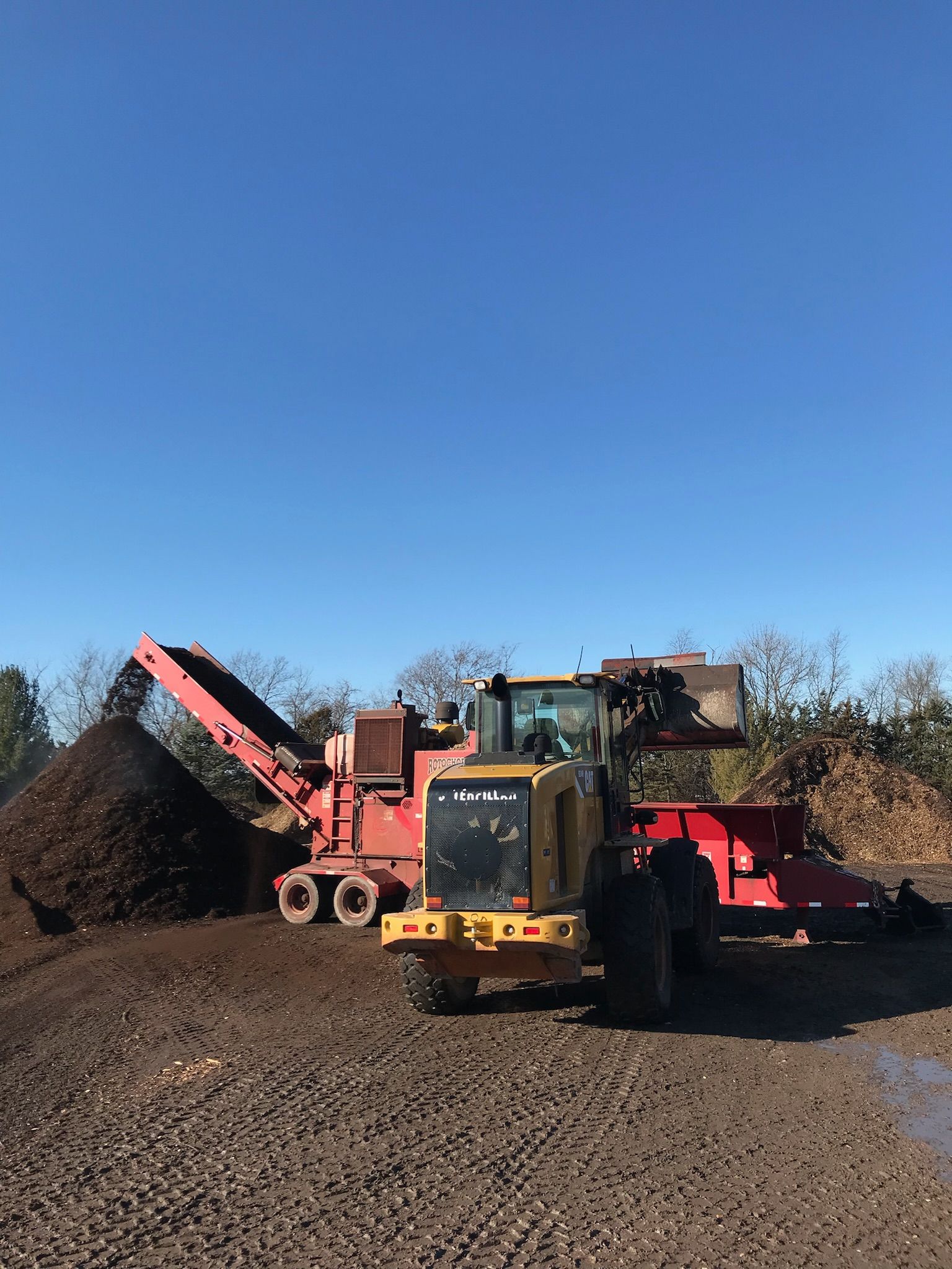 Red industrial wood chipper and yellow loader working outdoors on a brown dirt pile under a blue sky.