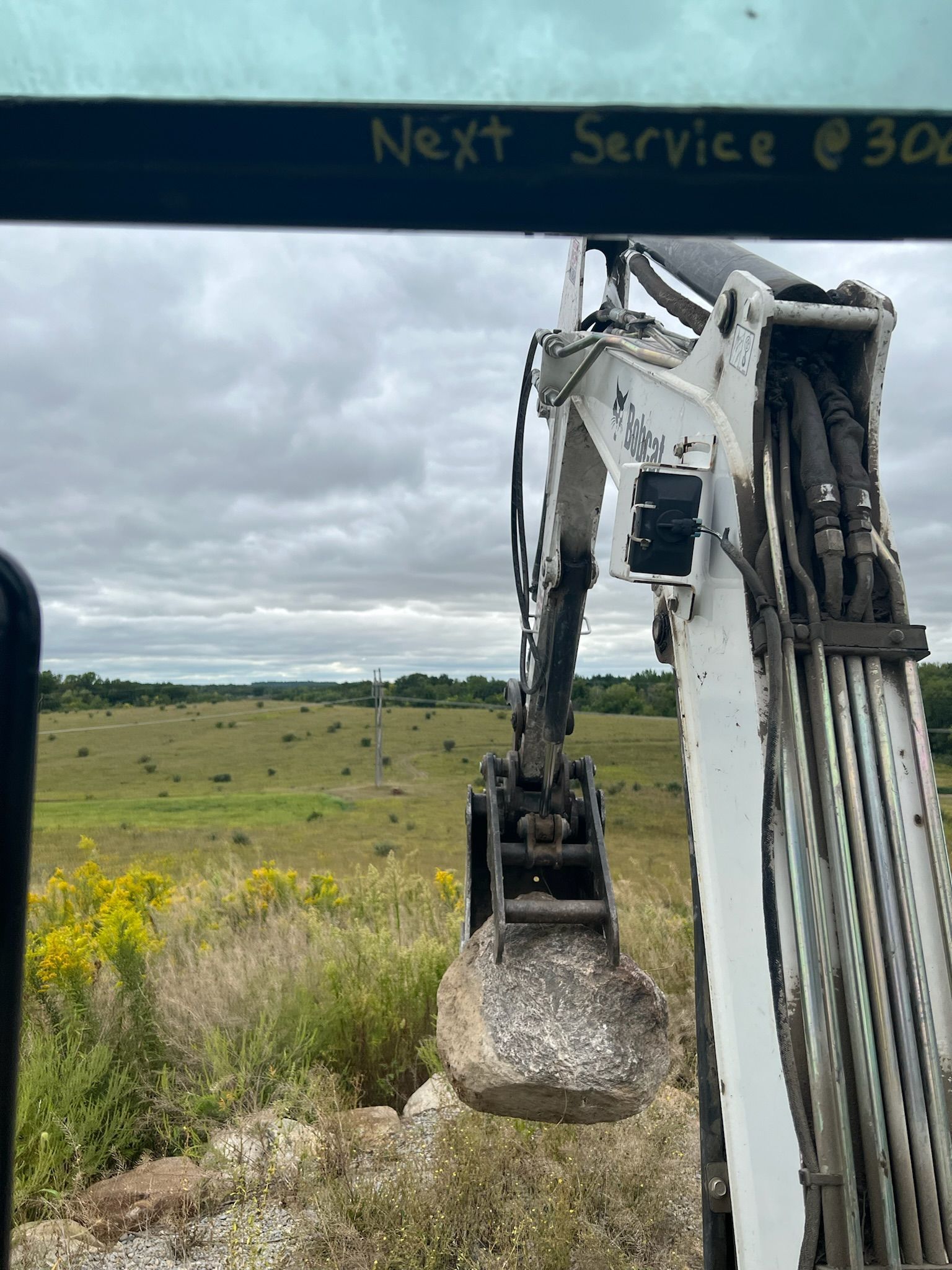 View from inside a Bobcat excavator cab, holding a rock in the bucket, overlooking a field.