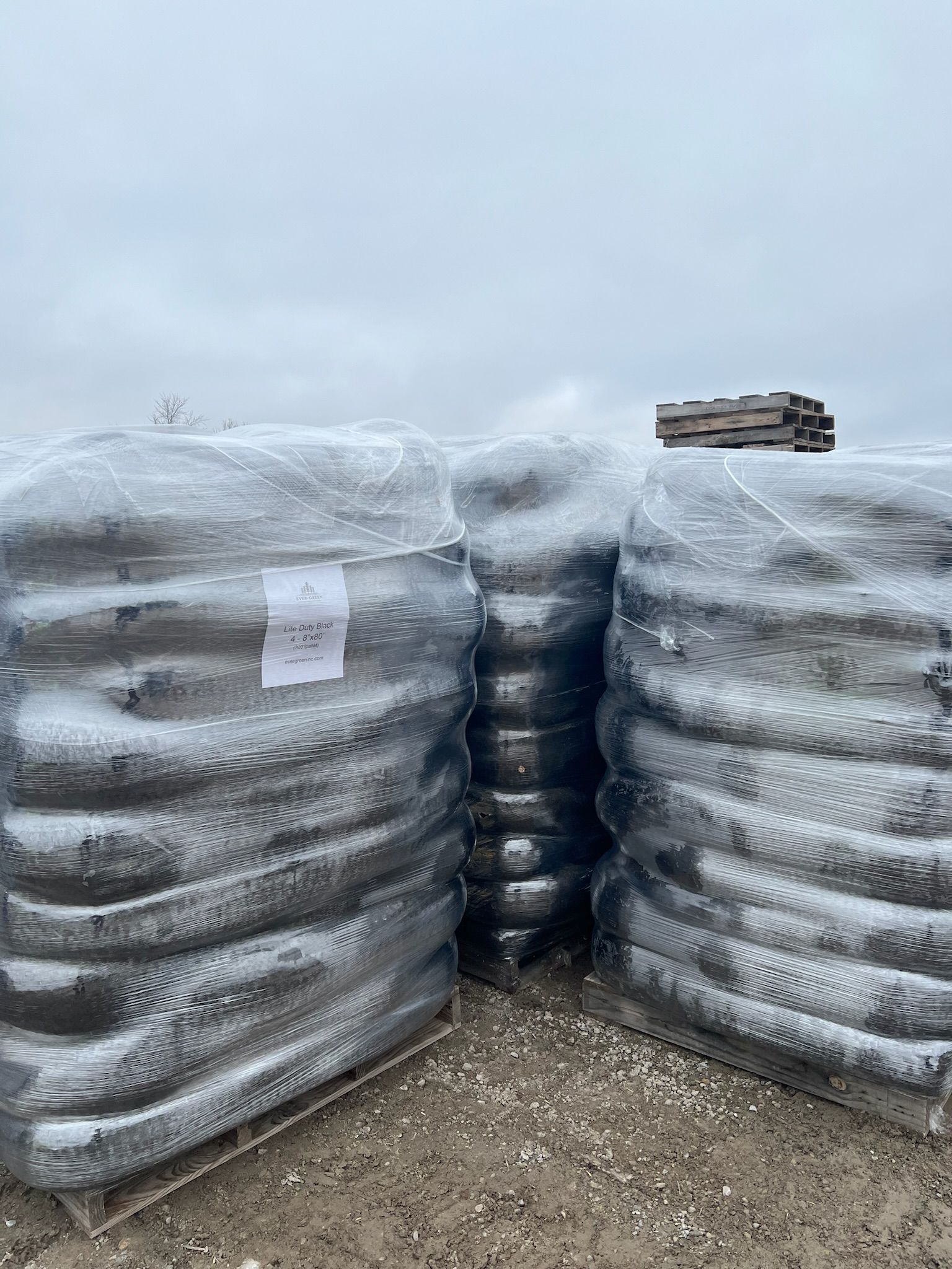 Three stacks of black bags wrapped in clear plastic, sitting on pallets outdoors under a cloudy sky.
