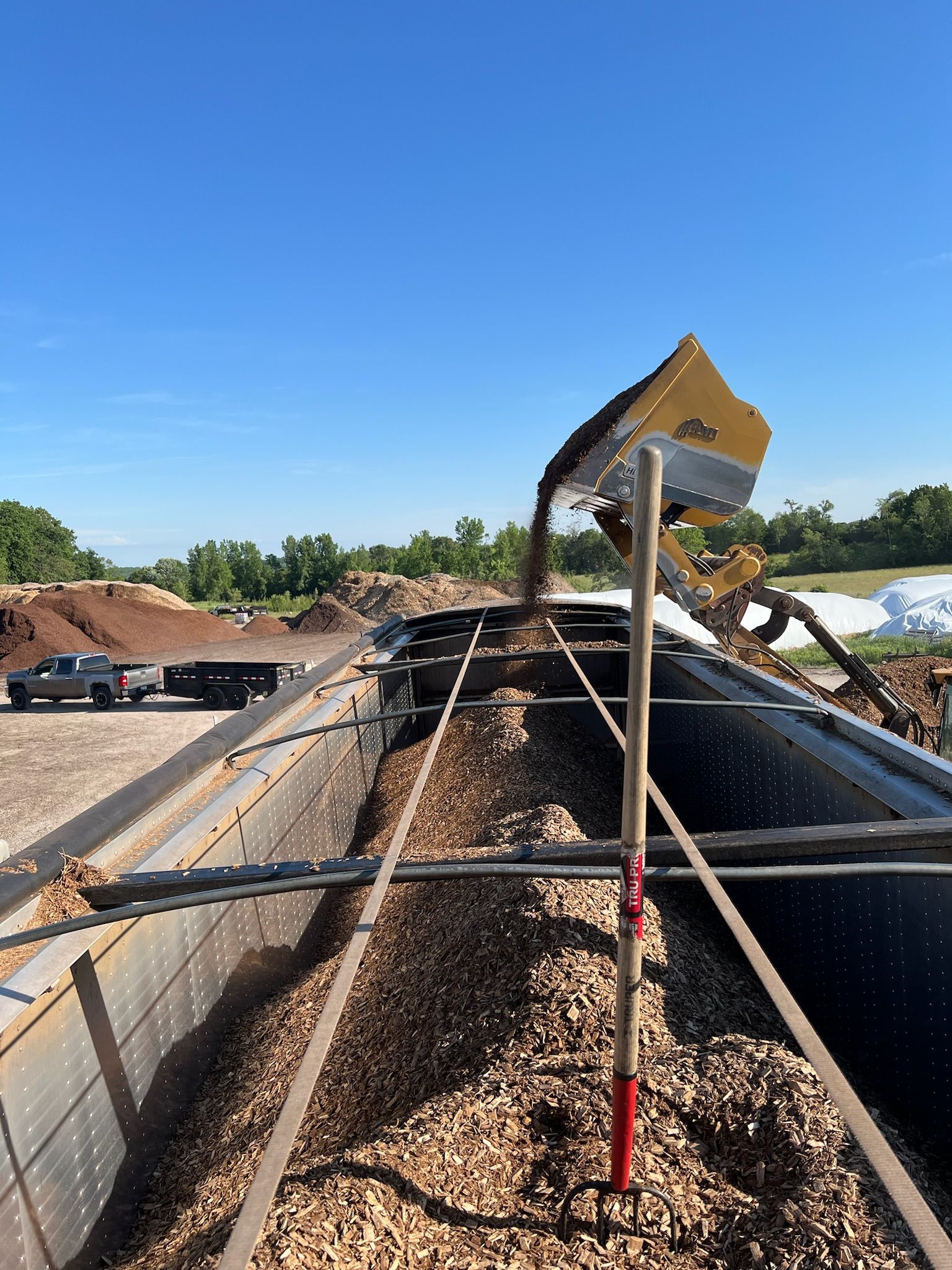 Wood chips being loaded into a long metal container by a yellow machine under a blue sky.