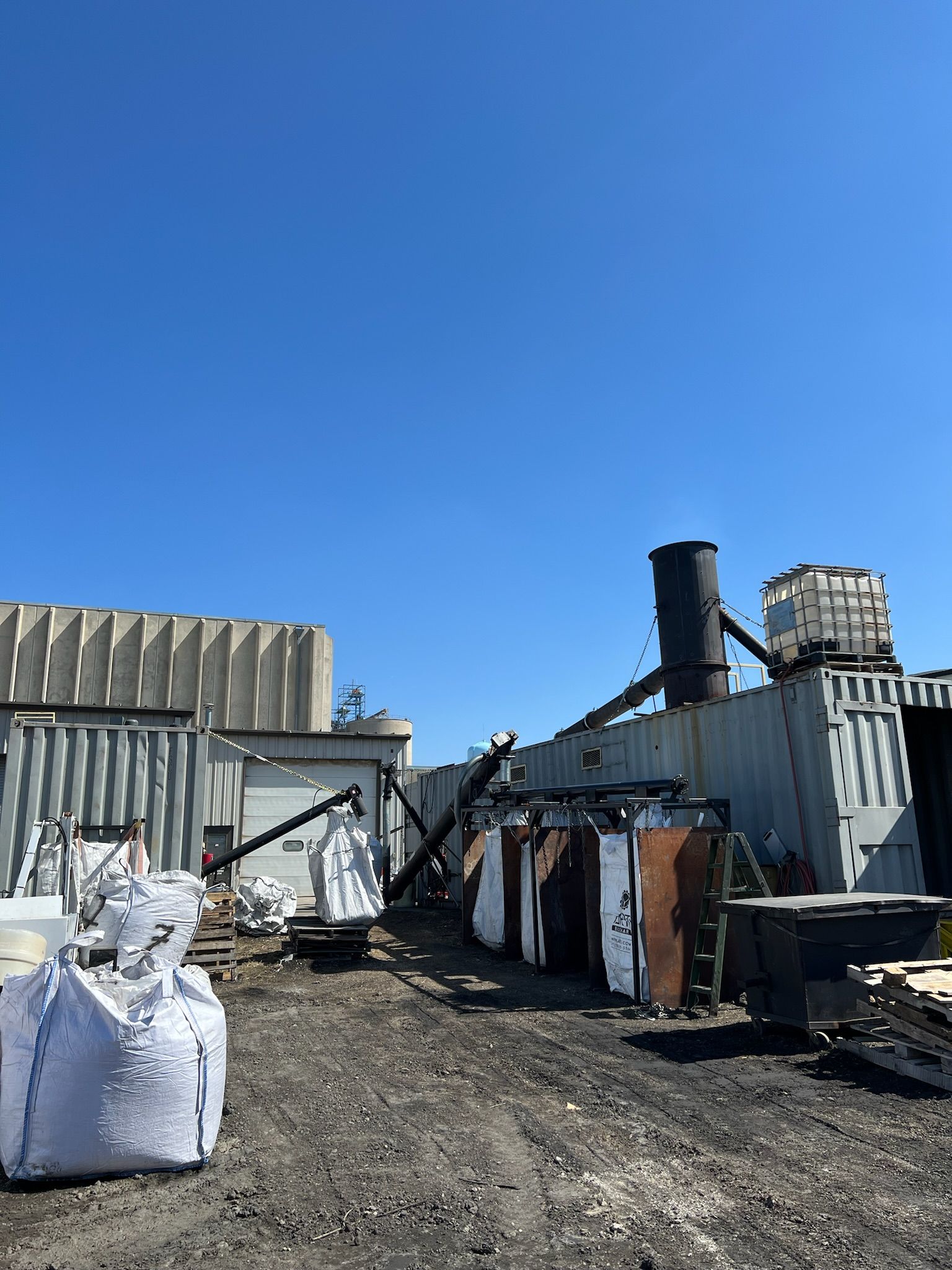 A yard with industrial equipment, containers, and a tall smokestack against a blue sky.