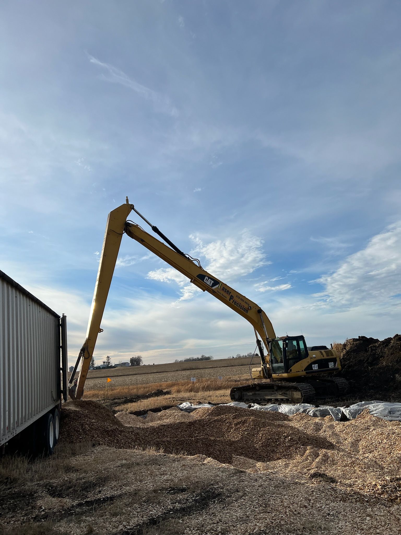 Excavator with an extended arm, working near a trailer, under a partly cloudy sky.