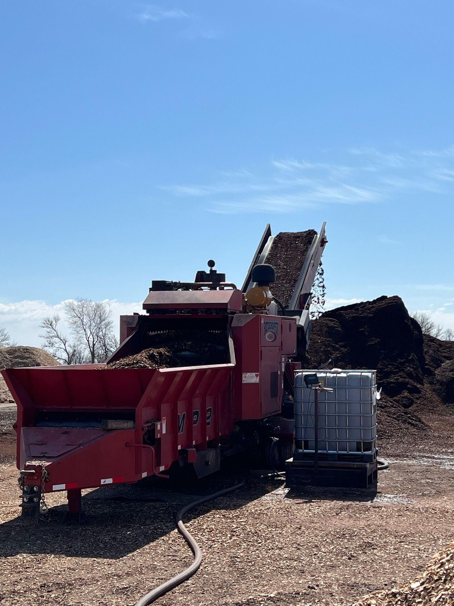Red wood chipper processing wood chips, with a pile of chips, and a water tank, against a blue sky.