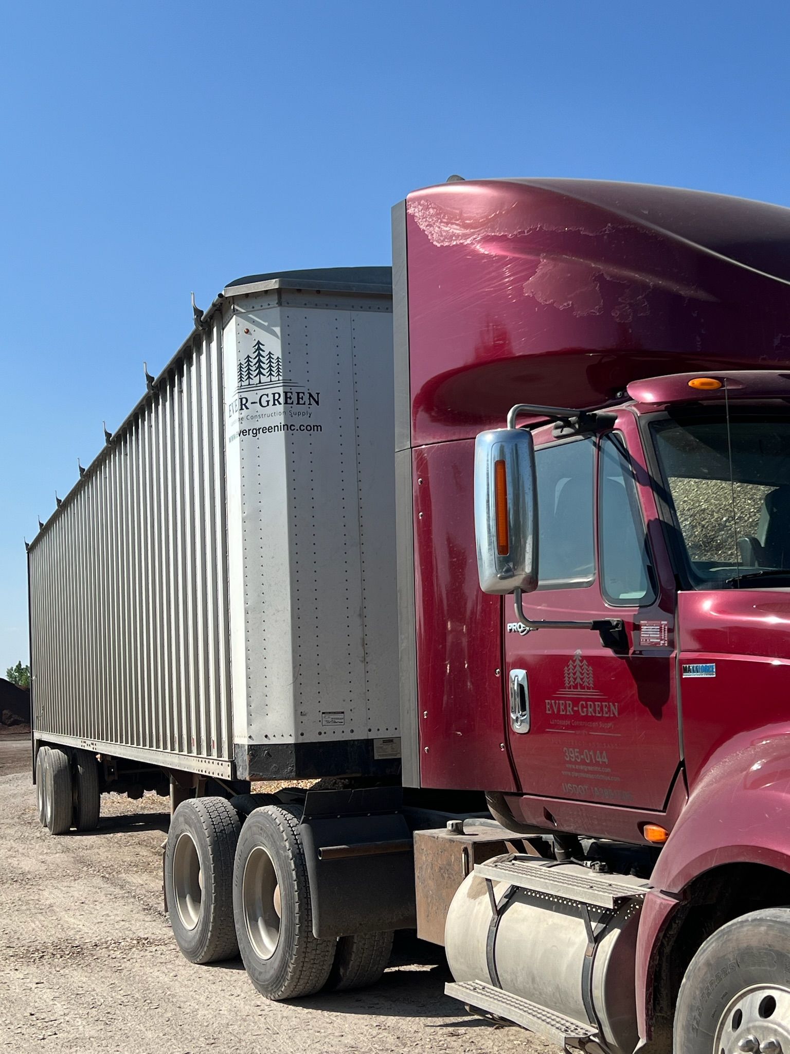 Red semi-truck hitched to a white trailer on a dirt road, under a clear blue sky.