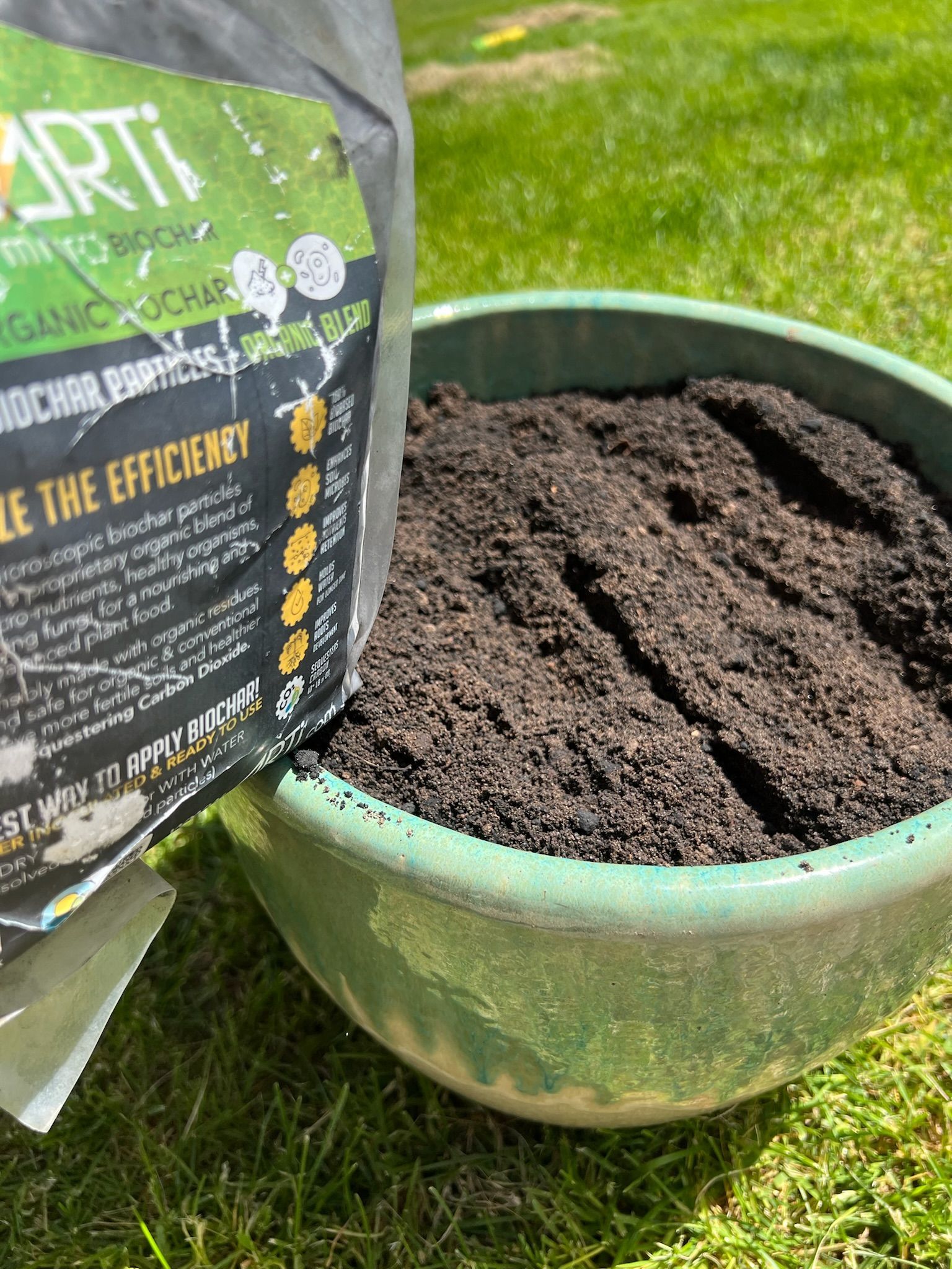 Dark soil being poured from a bag into a green pot, set on grass.