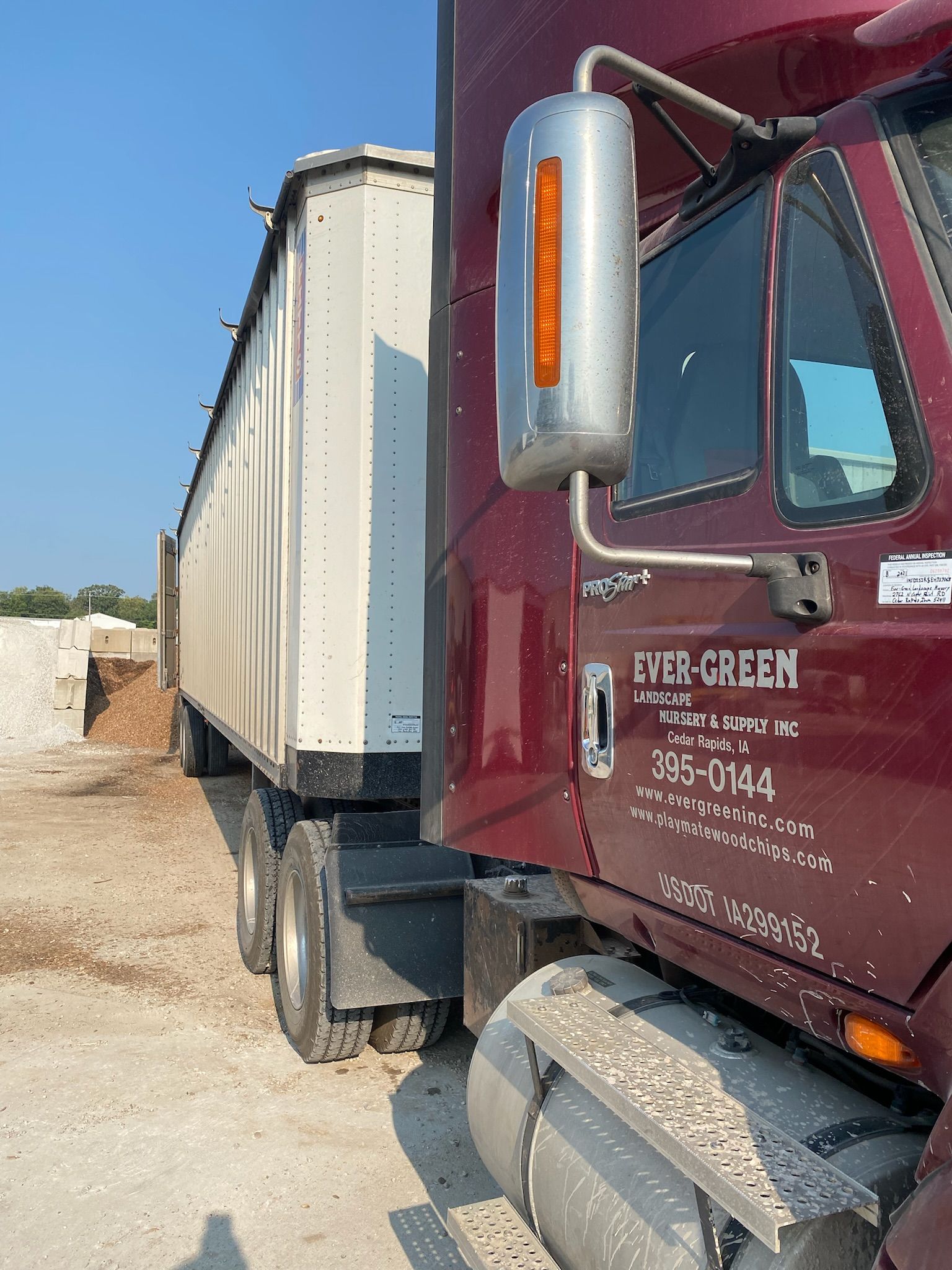 Red semi-truck with white trailer parked on gravel, under a blue sky. 