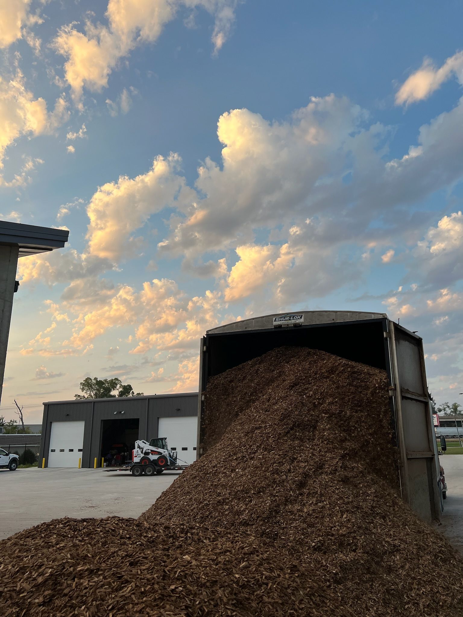 Truck bed spilling mulch in front of a building with a cloudy sky backdrop.