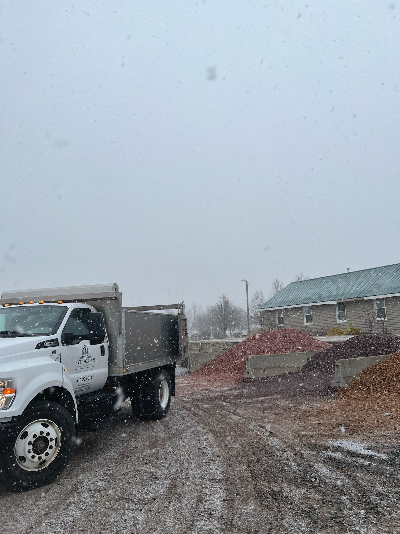 White dump truck on gravel driveway next to piles of mulch, with a building in the background. Snow falling.