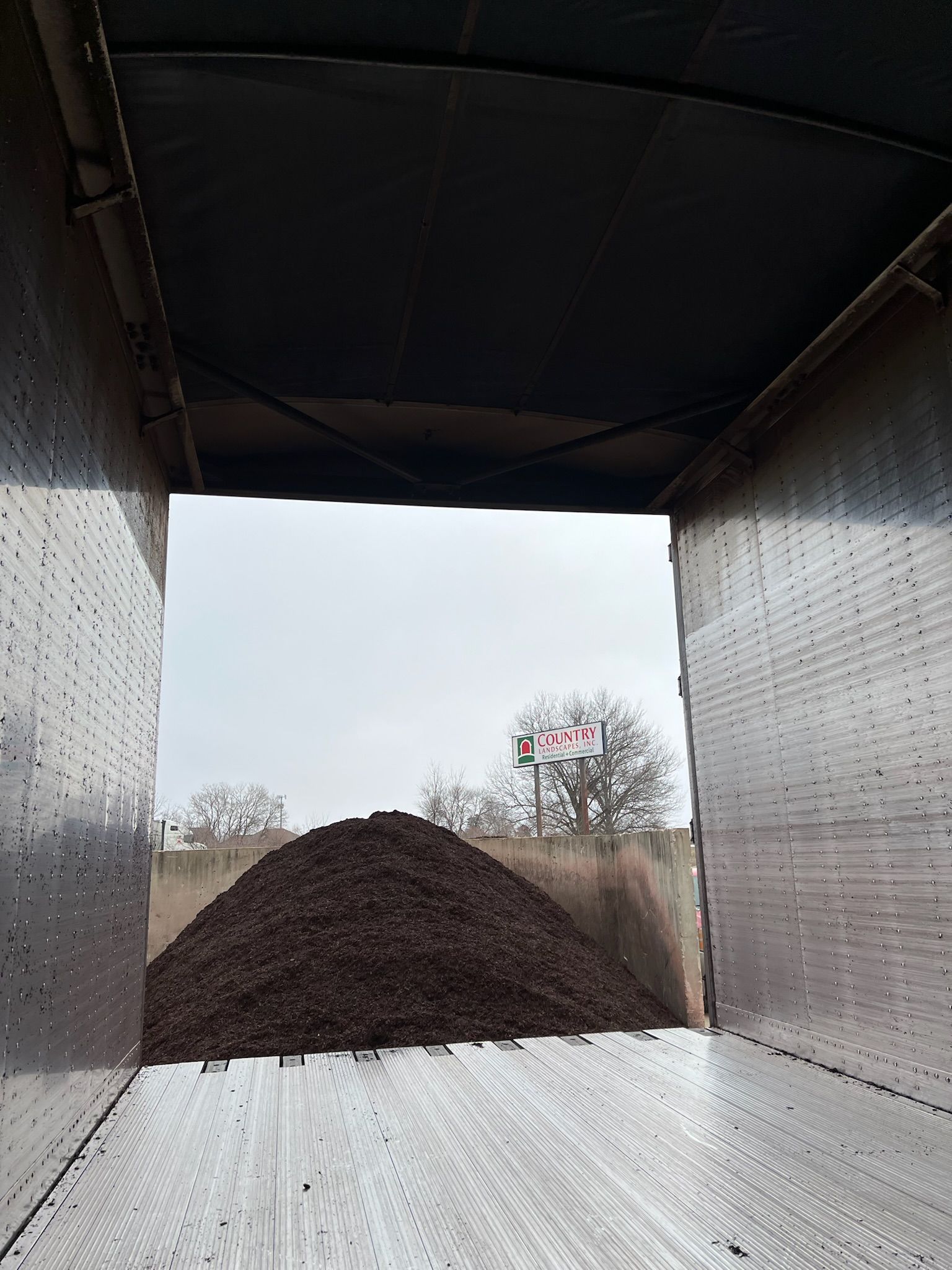 View from inside a trailer filled with dark brown material, looking out at a cloudy sky and distant buildings.