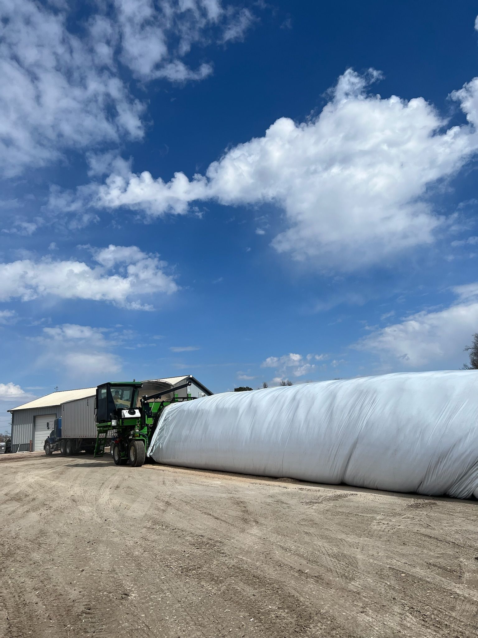 Green tractor filling a large white silage bag with feed on a farm under a blue sky.