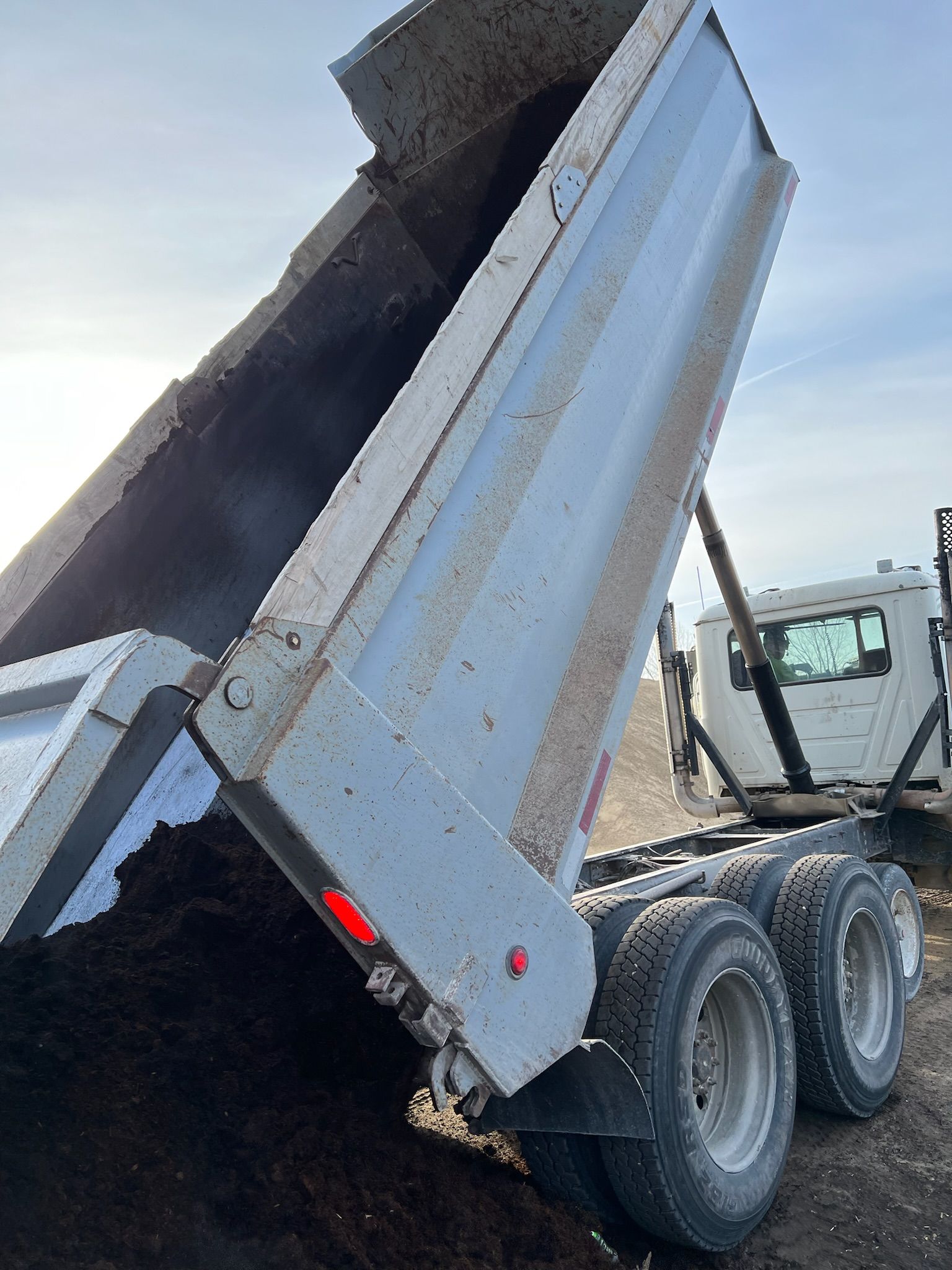 Dump truck bed raised, dumping dark material. Light sky, truck tires, and cab visible.