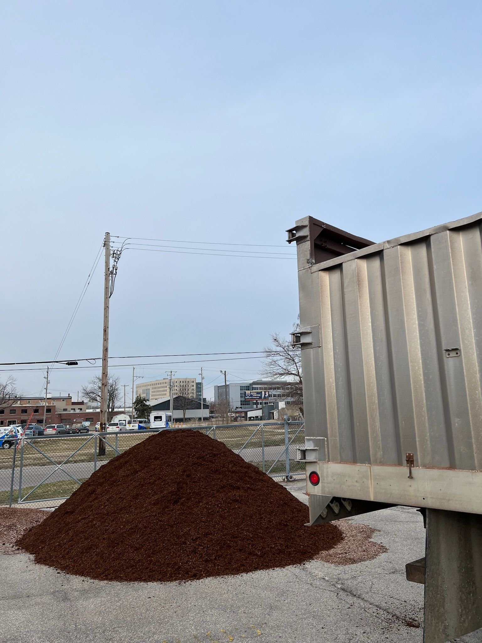 Dump truck dumping a pile of brown mulch on a gravel lot under a cloudy sky.