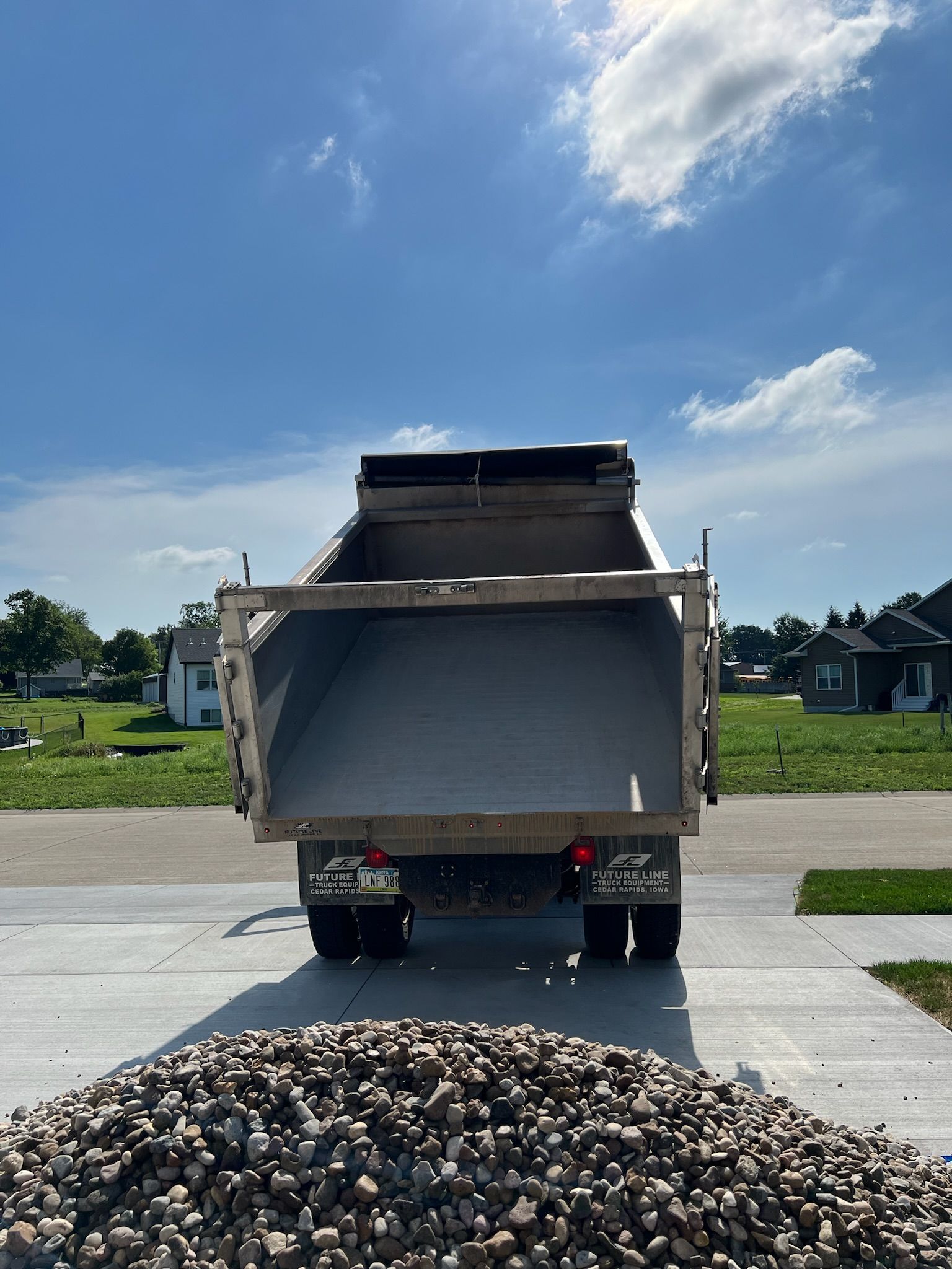 Dump truck bed filled with gravel, backed onto a driveway on a sunny day.