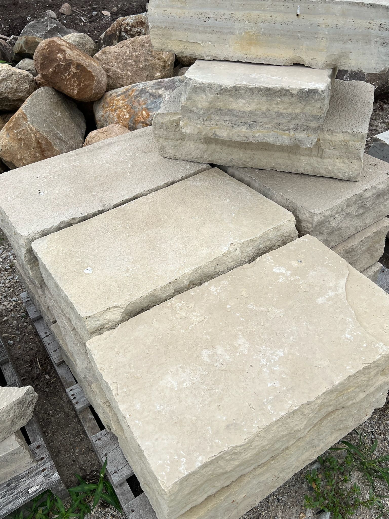 Stacked rectangular light-colored stone blocks, some with rough edges, near larger rocks.