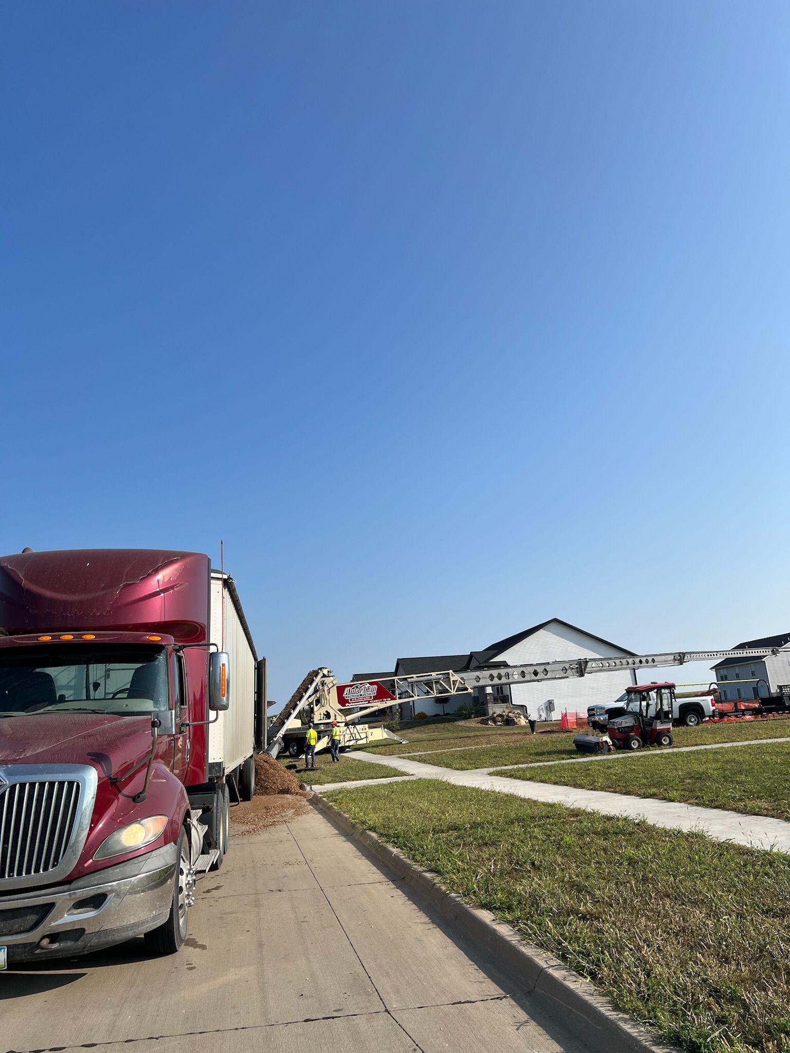 Red semi-truck parked on roadside. Construction site with buildings and equipment in the background under blue sky.
