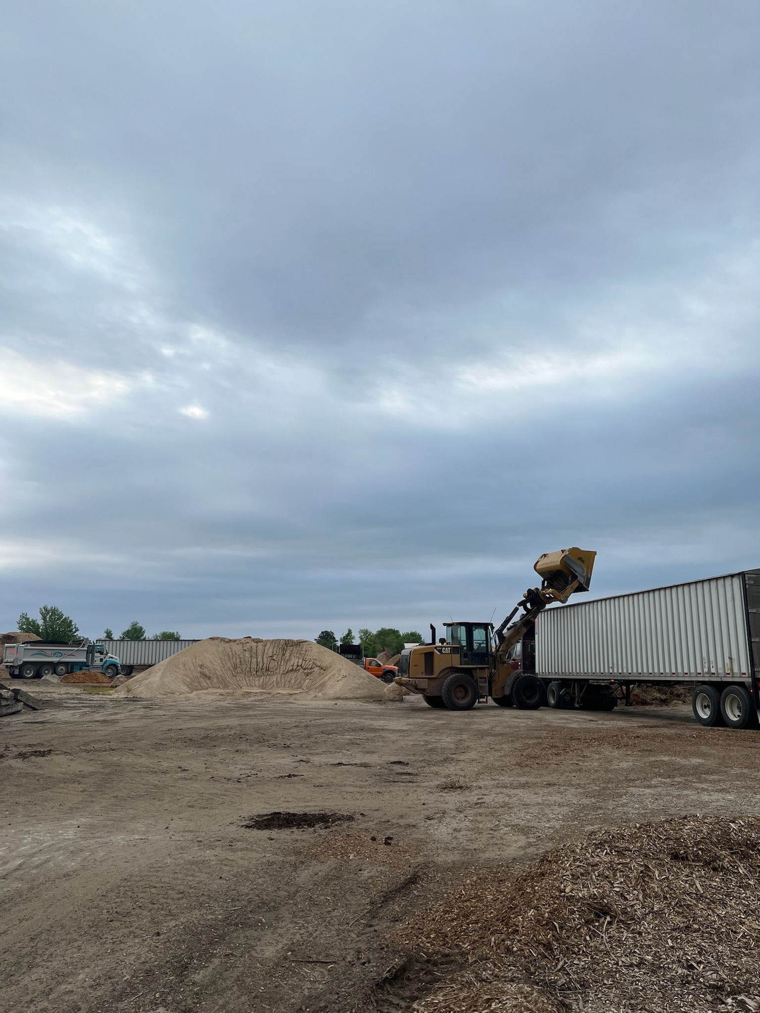 Front loader dumping wood chips into a semi-trailer on a cloudy day in a lumberyard.