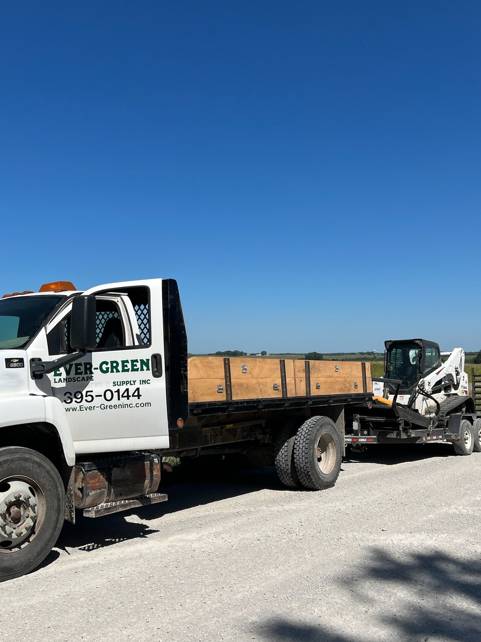 White truck with a flatbed trailer transporting a skid steer loader on a gravel road, under a blue sky.