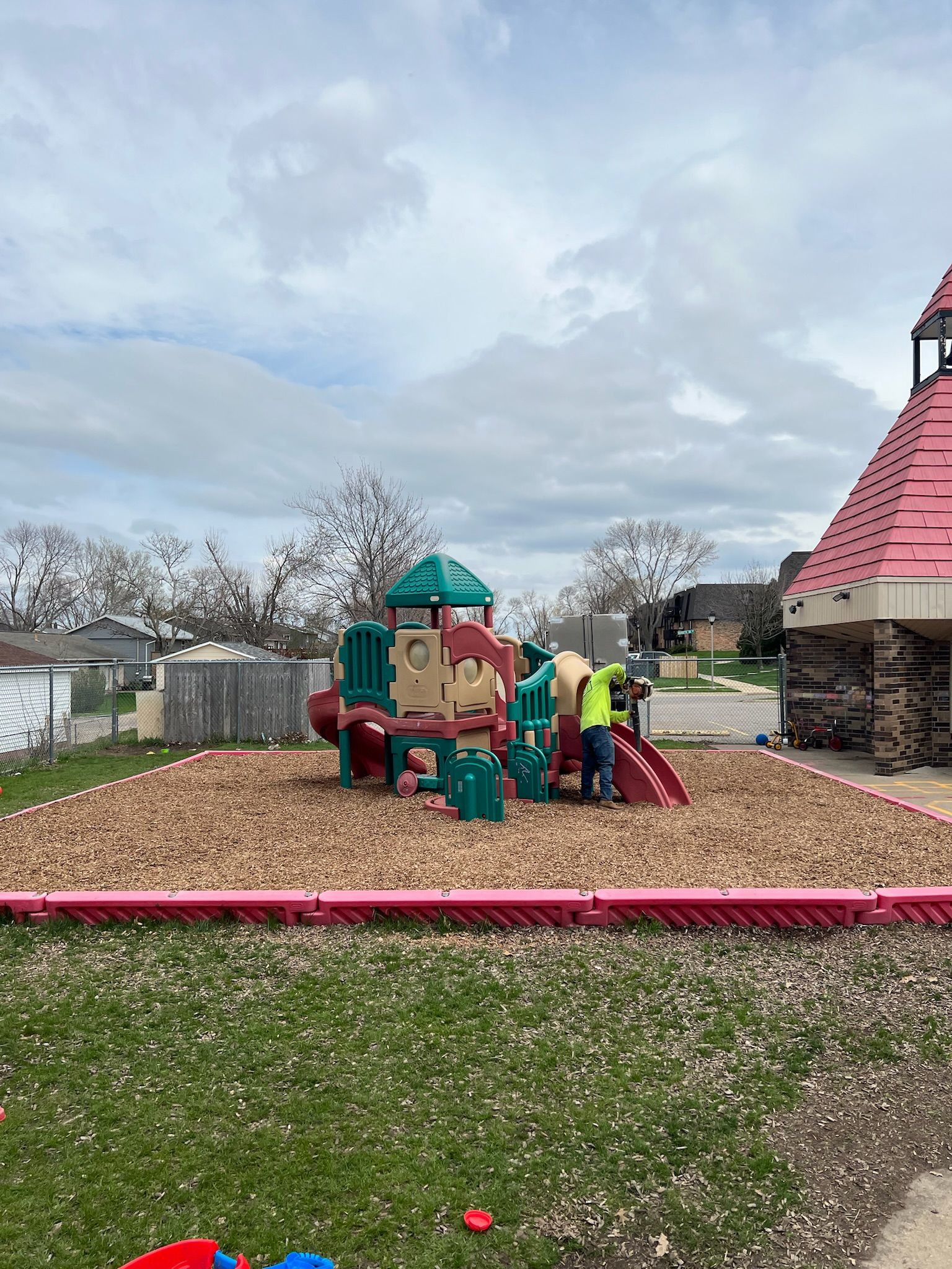 Playground with wood chips, red and green play structure, person adjusting slide, overcast sky.