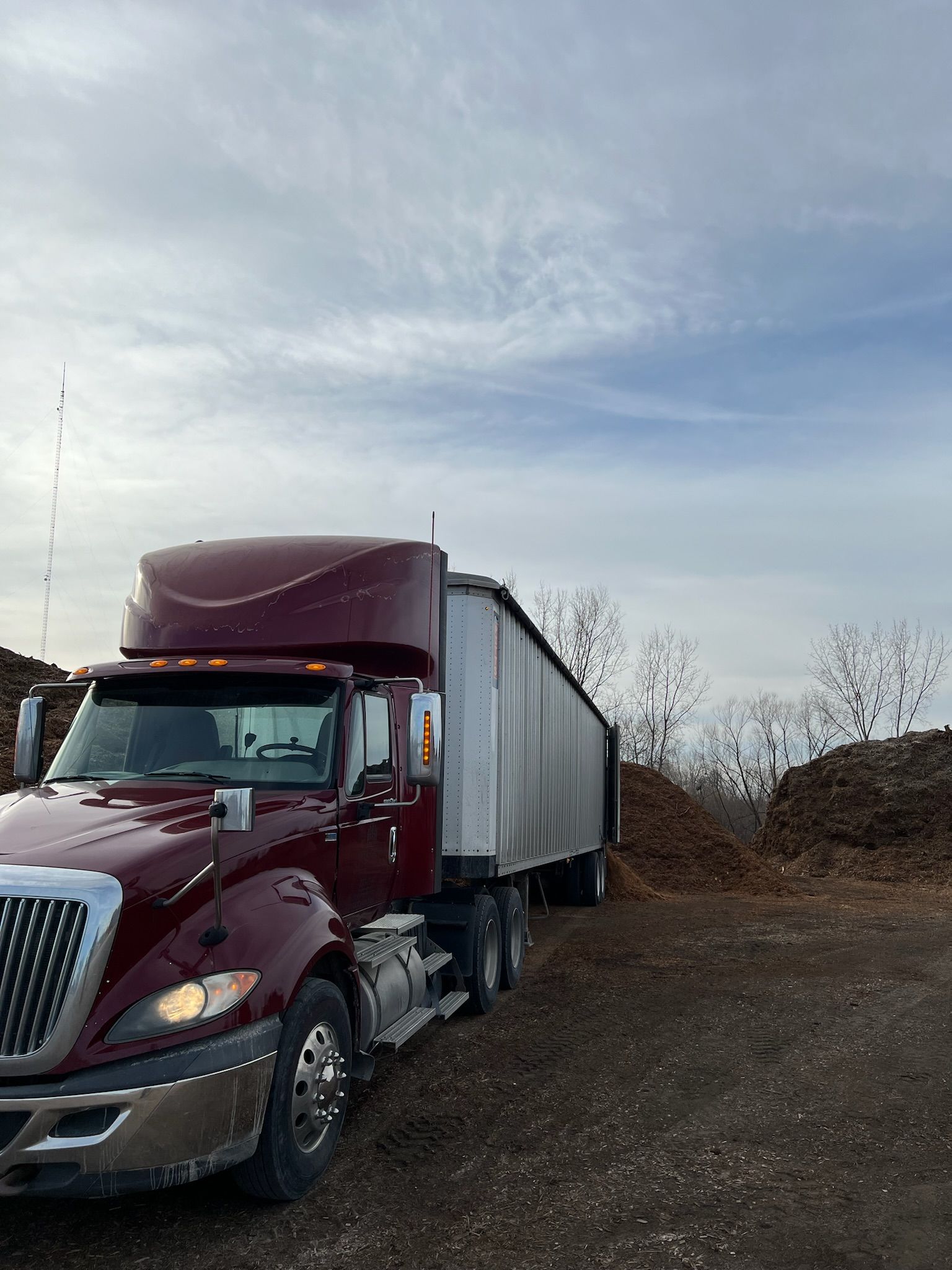 Maroon semi-truck with trailer on a dirt lot, near piles of material under a cloudy sky.