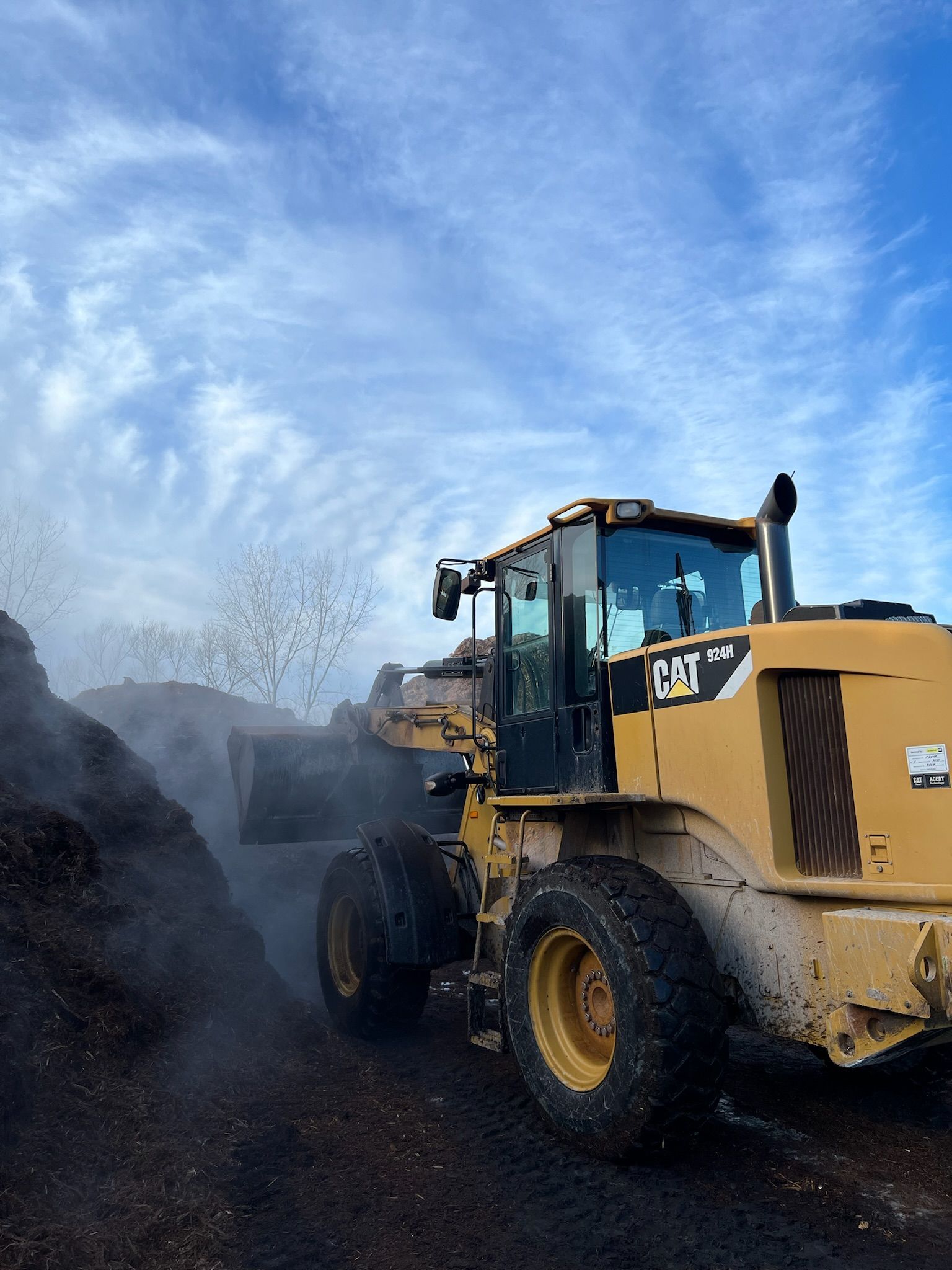 Yellow Caterpillar loader moving dark mulch under a cloudy blue sky.