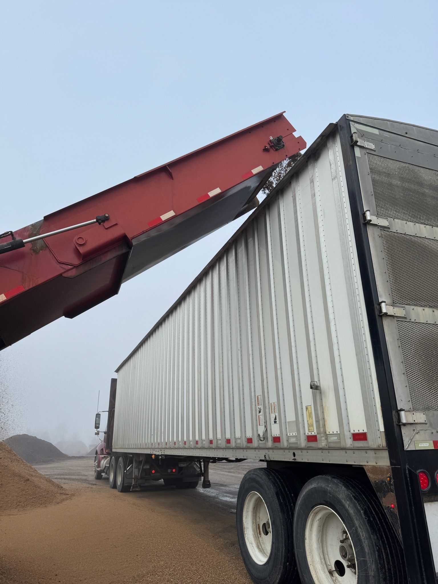A semi-truck being loaded with brown material by a red conveyor belt; outdoors on an overcast day.