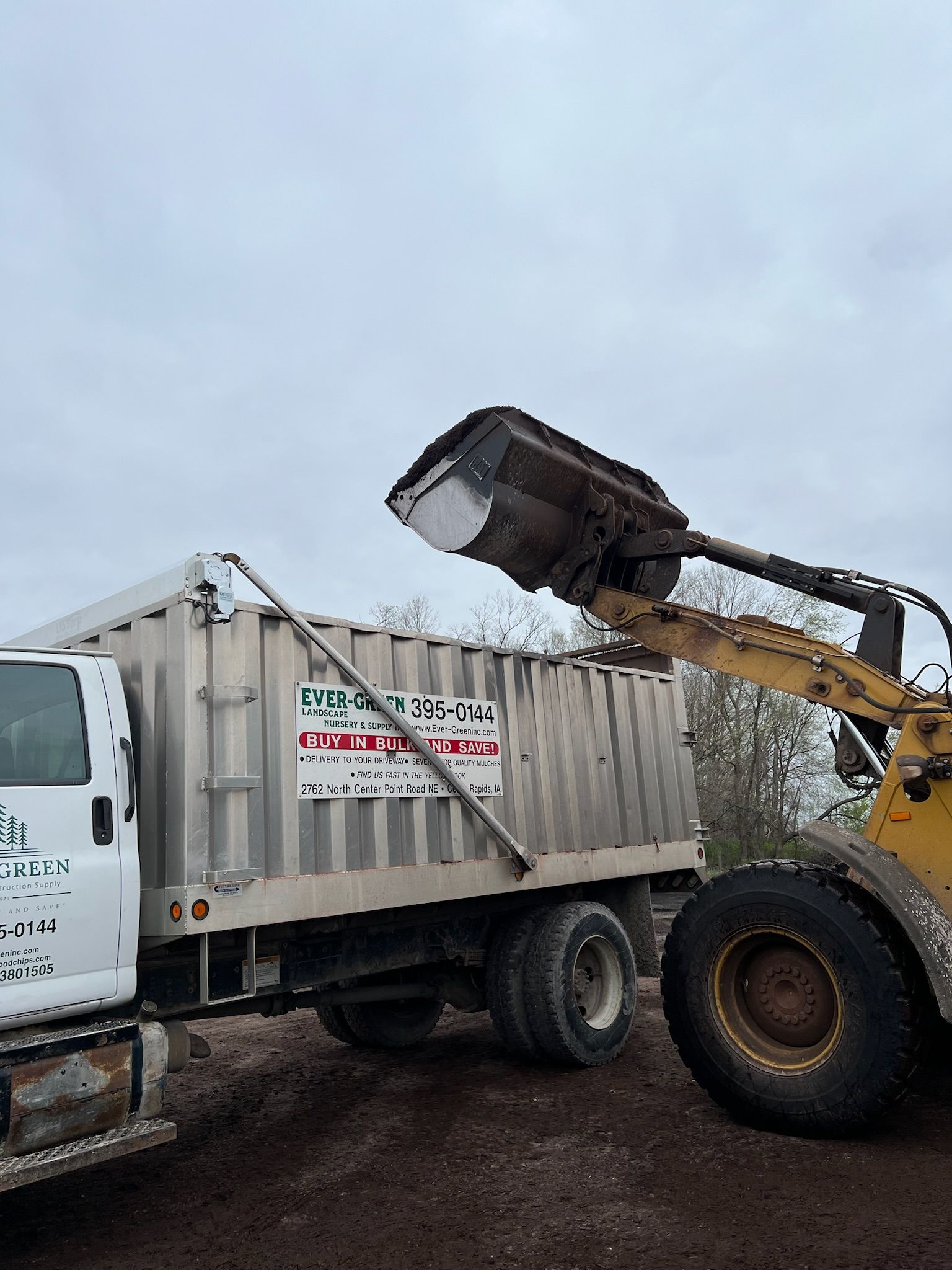 A front-end loader is dumping material into the bed of a dump truck on a brown surface. Cloudy sky.