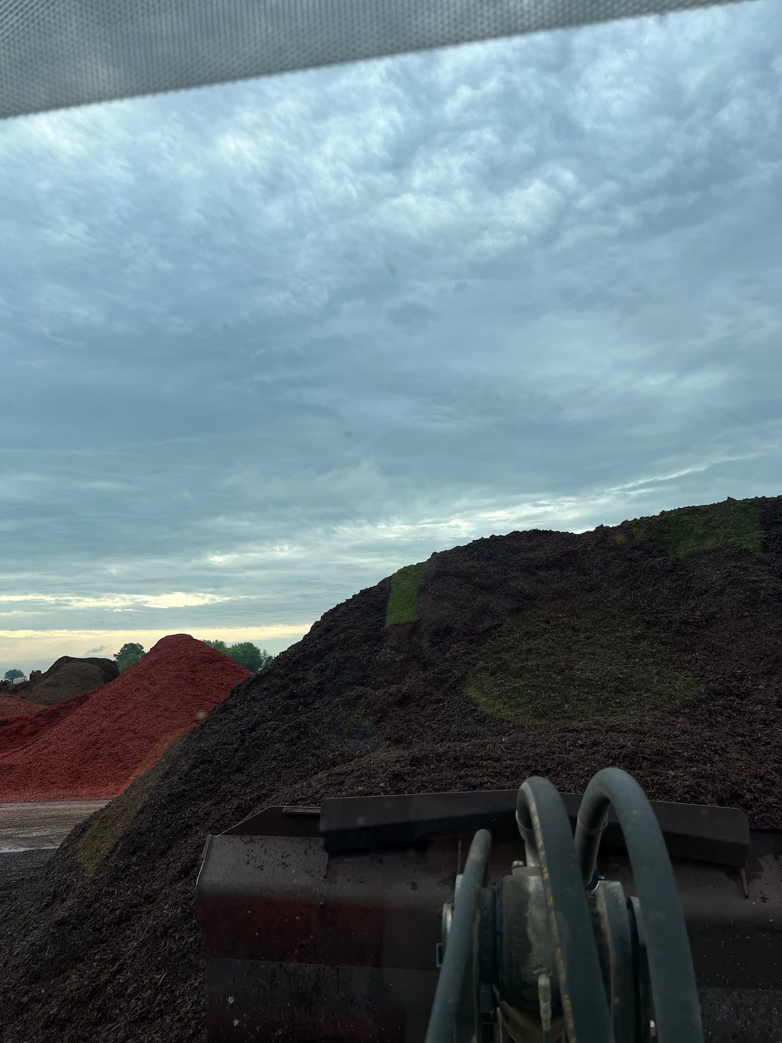 Large piles of dark brown material under a cloudy sky, with a red pile visible on the left and a machine bucket in foreground.