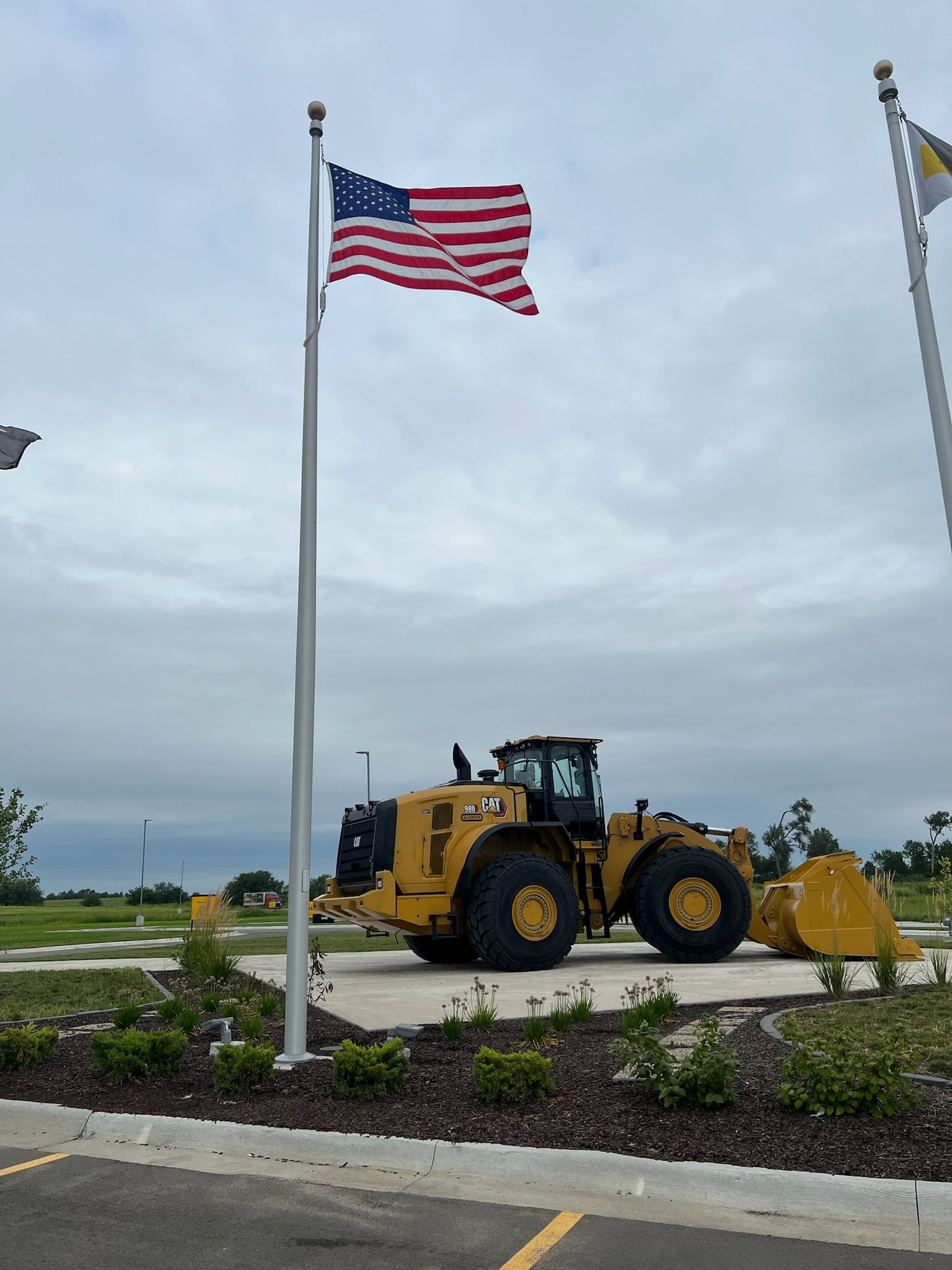American flag on pole with yellow construction vehicle on a concrete slab, overcast sky.